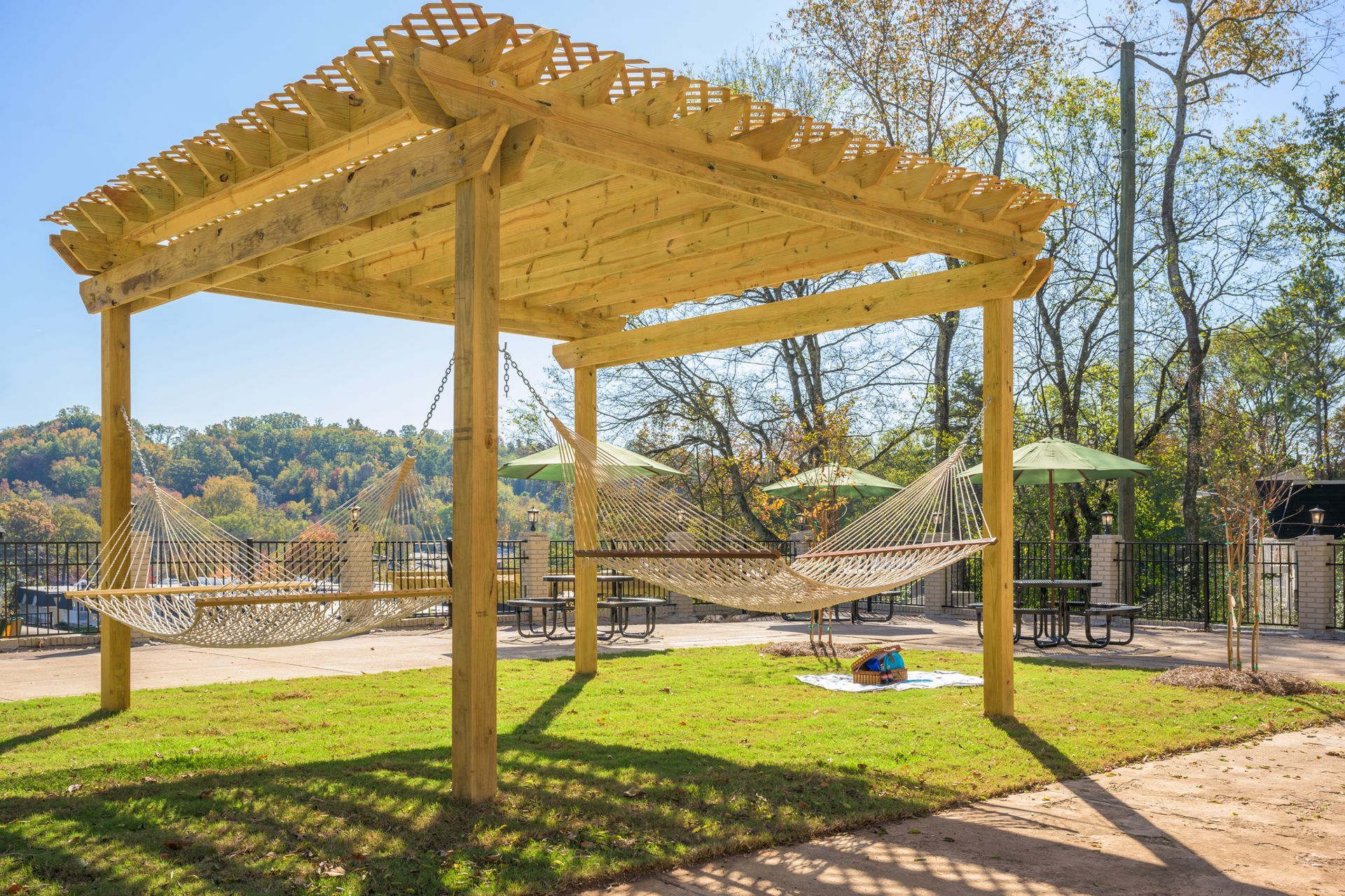 A wooden pergola stands on a lawn, featuring two white rope hammocks hanging between its posts under a clear blue sky.