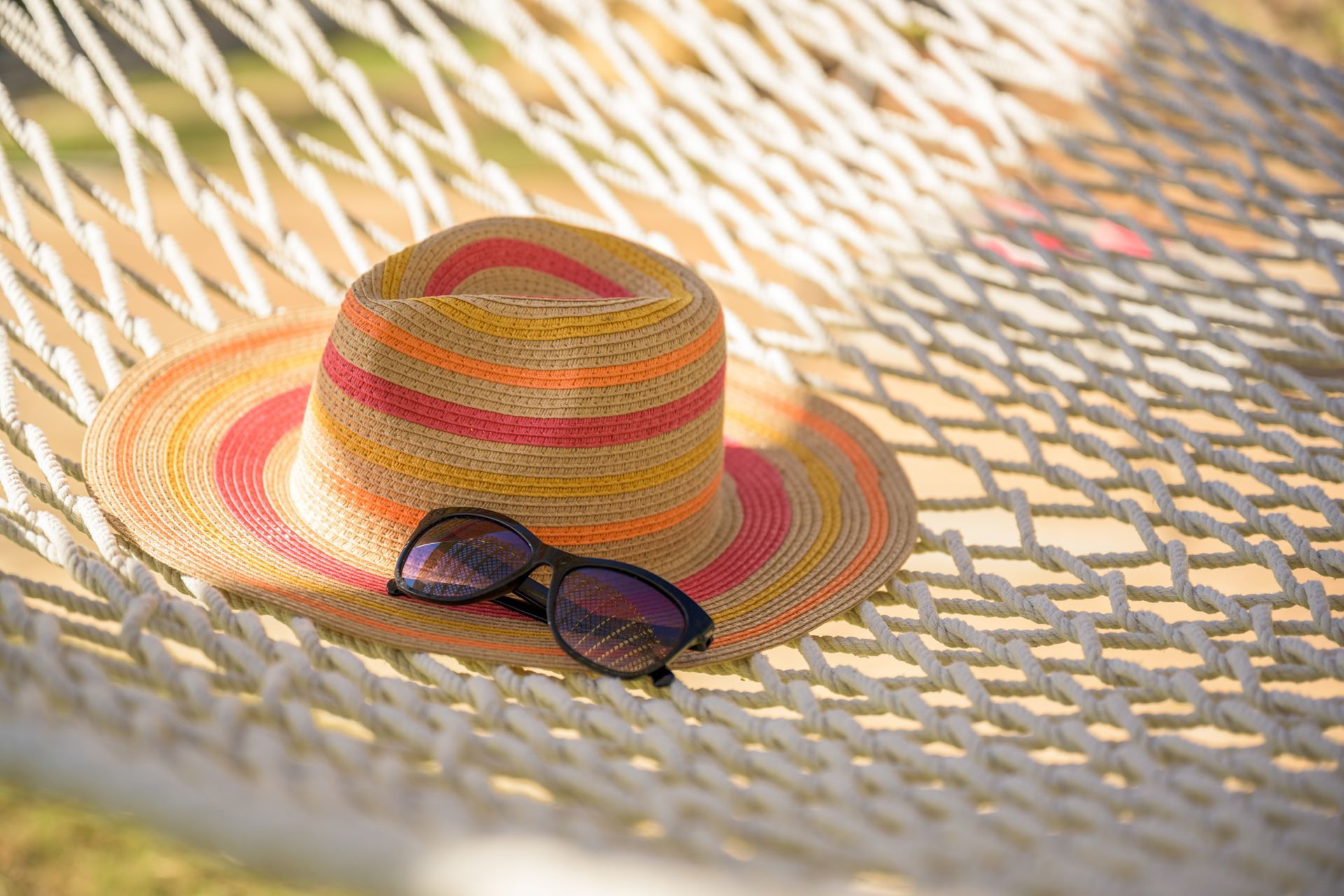 A sun hat with orange and pink stripes and black sunglasses resting on a white woven hammock.
