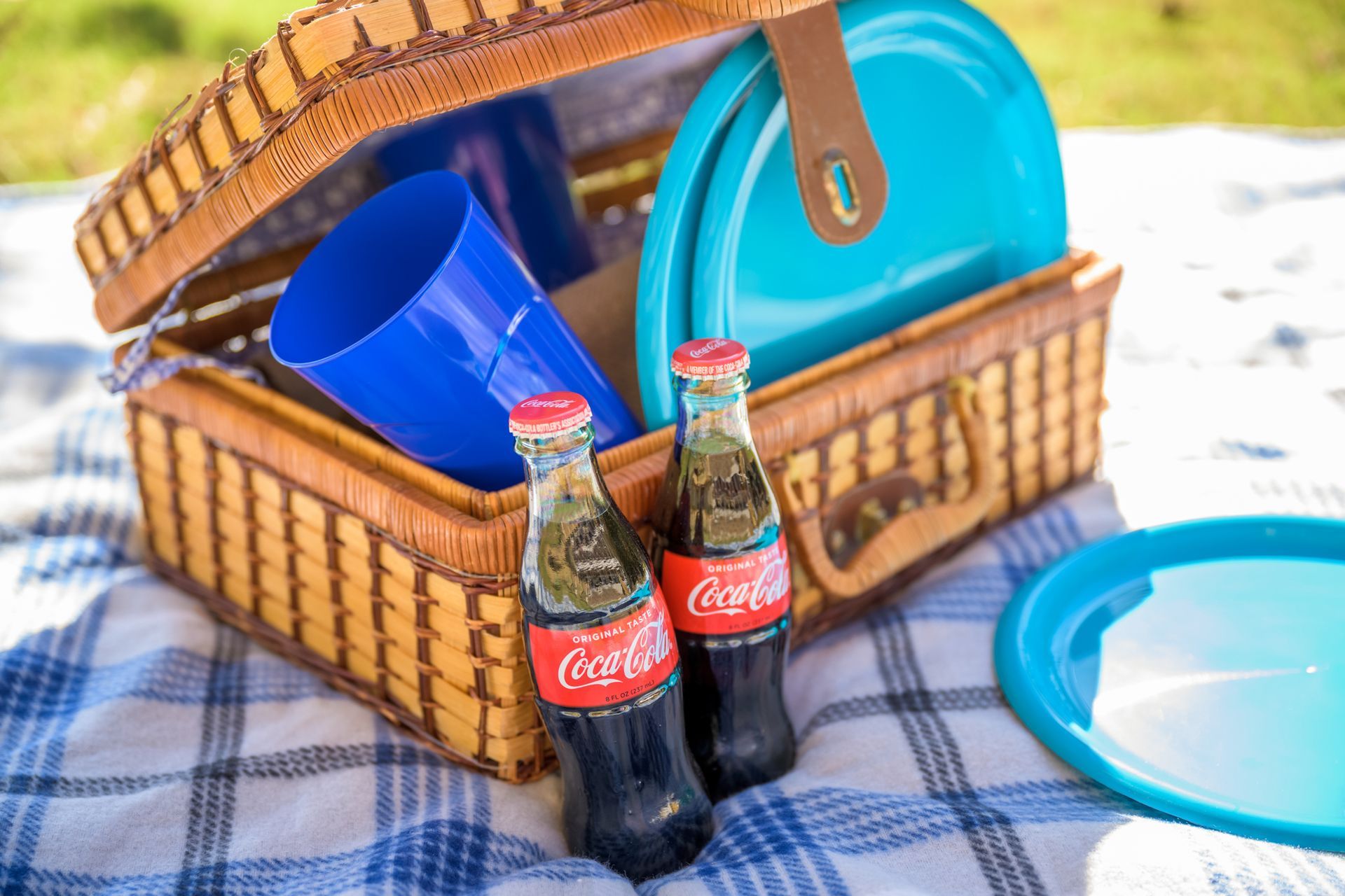 A picnic basket containing blue plates and cups sits on a plaid blanket with two glass bottles of Coca-Cola in front.