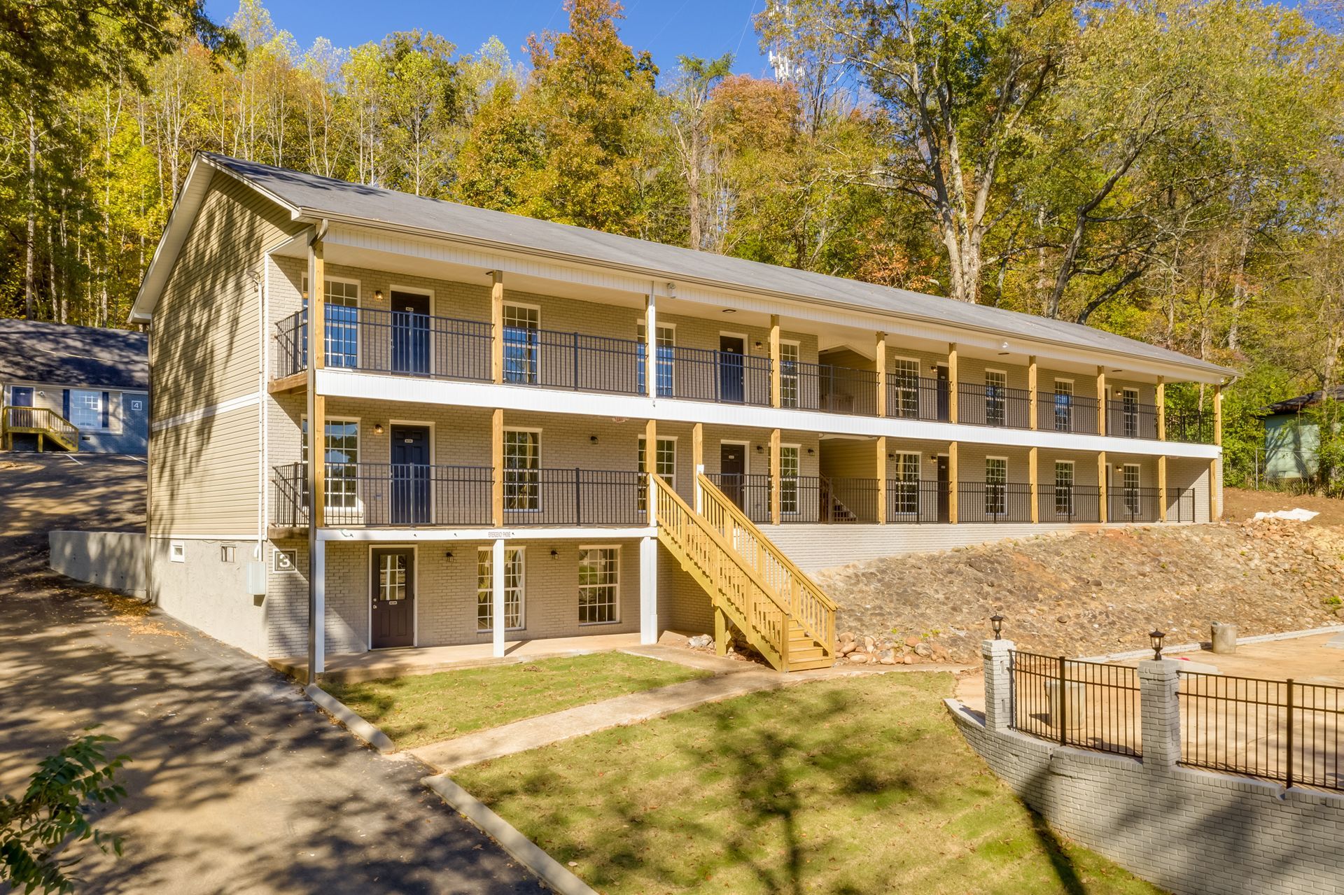 A three-story, light-colored motel building with outdoor balconies, wooden stairs, and a forest backdrop.