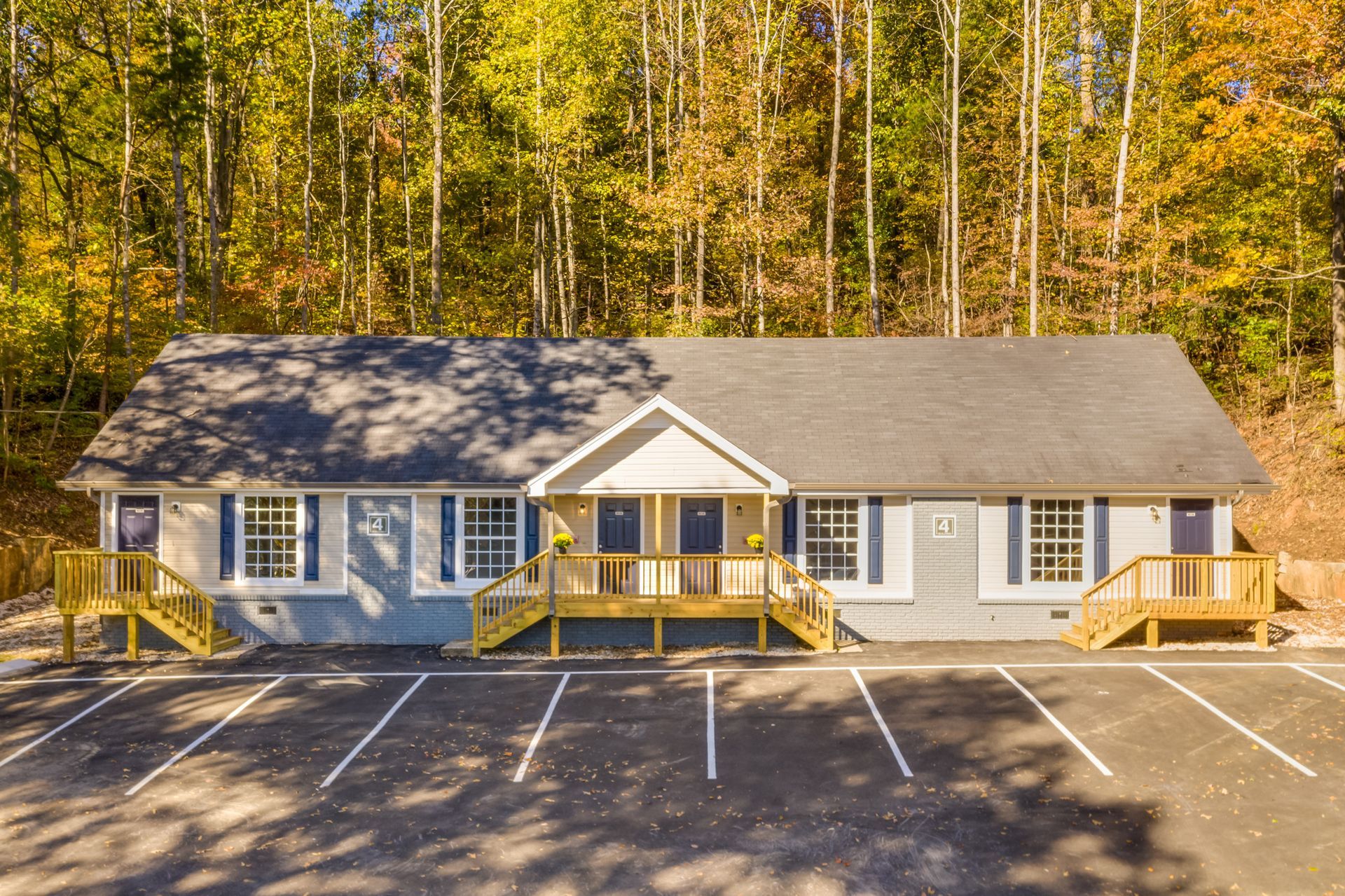 Single-story building with blue shutters and wooden porches, fronted by a parking lot against a backdrop of autumn trees.