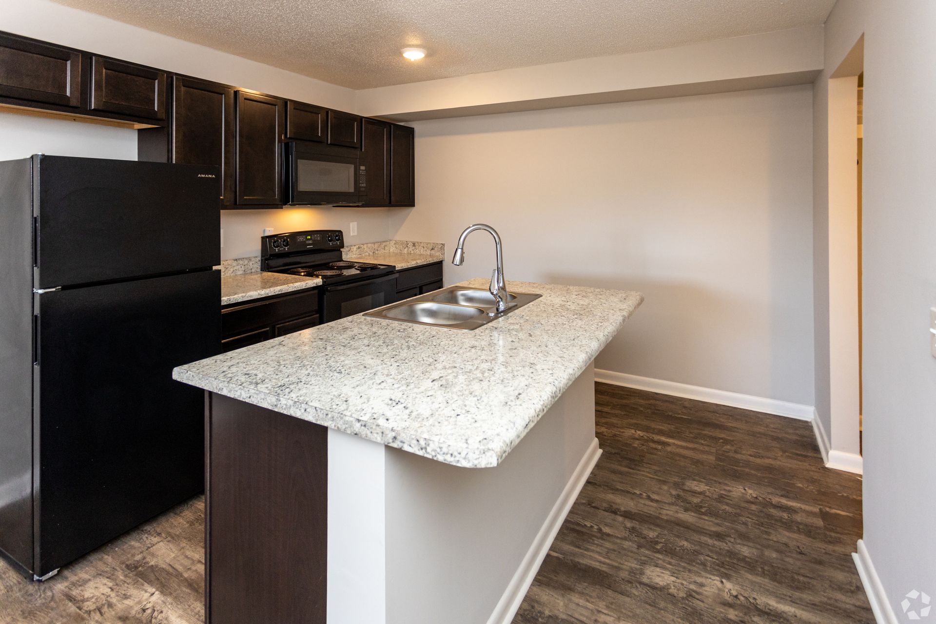 A modern kitchen with dark wood cabinets, black appliances, light granite countertops, and wood-style flooring.