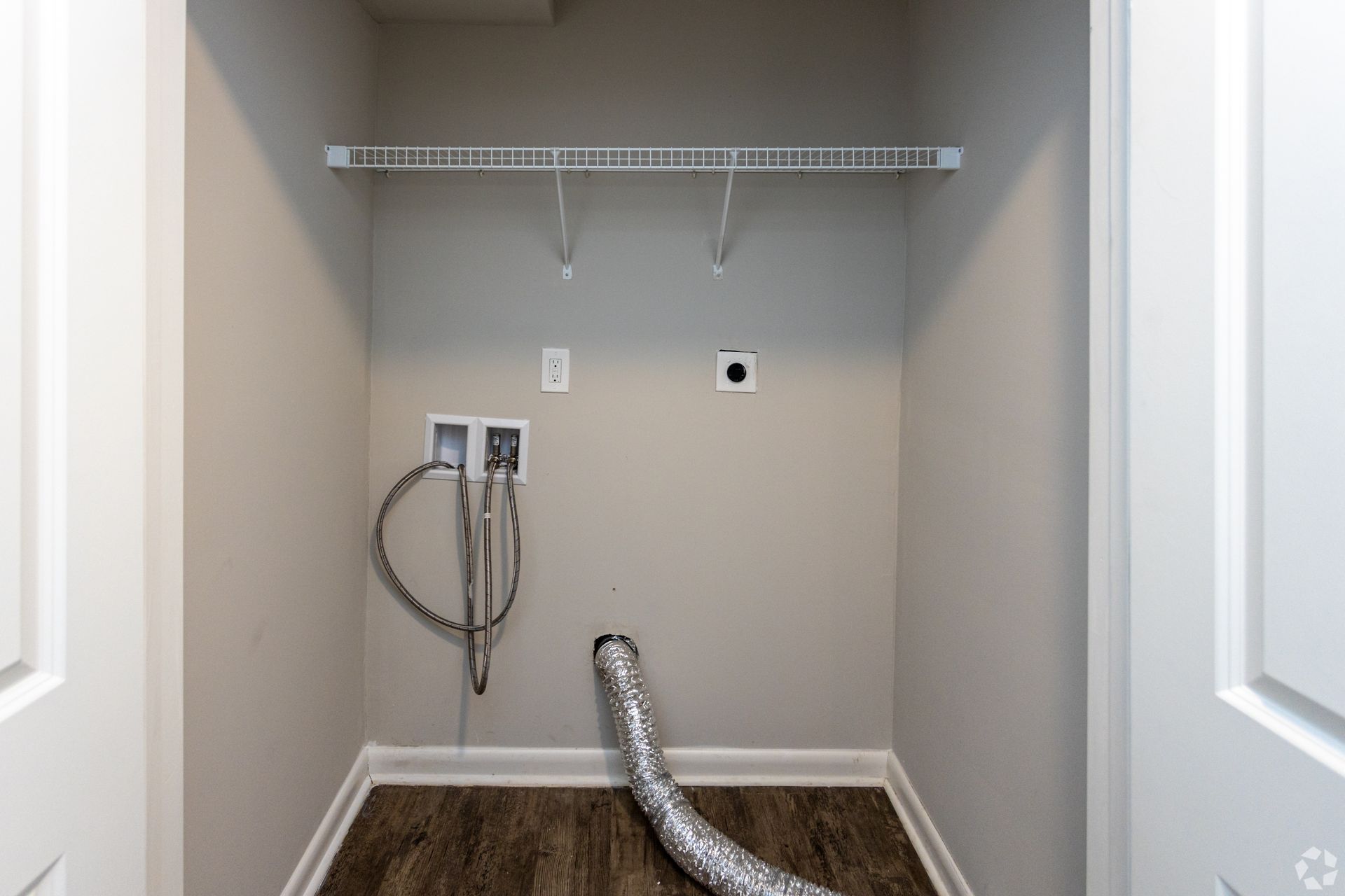 A small, empty laundry closet with light gray walls, wood floors, a wire shelf, and hookups for a washer and dryer.