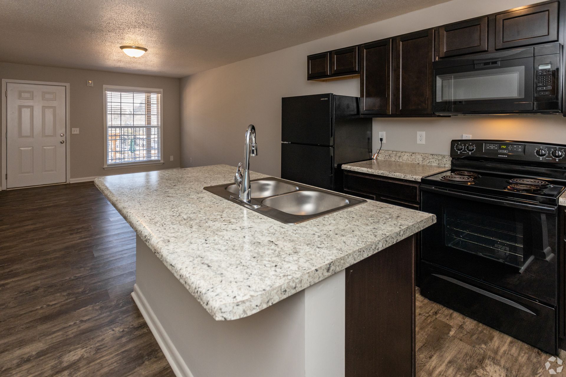 A kitchen with a granite-topped island, black appliances, and dark wooden cabinets in an open-concept apartment.