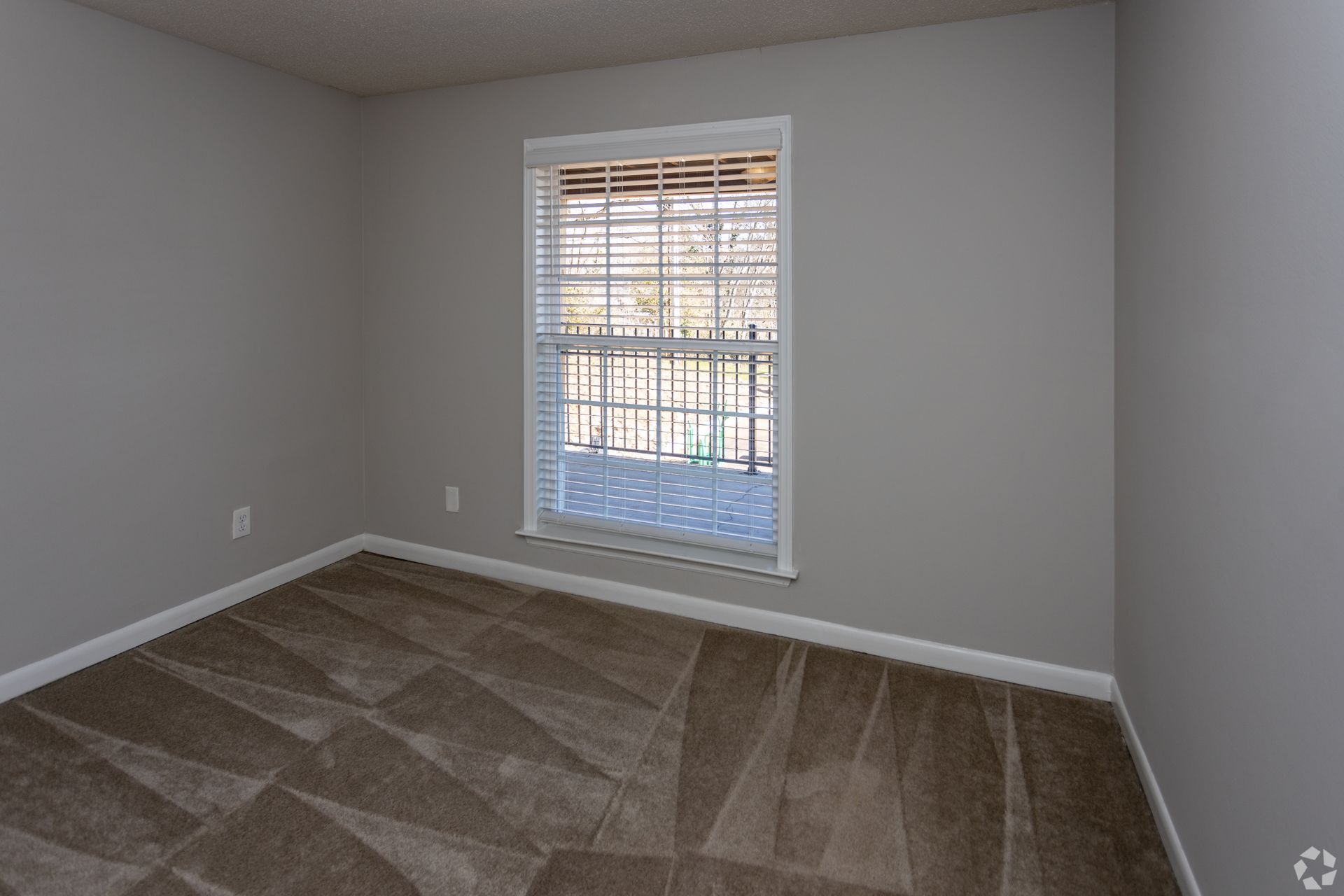 An empty bedroom with gray walls, brown patterned carpet, and a single window overlooking an outdoor balcony.
