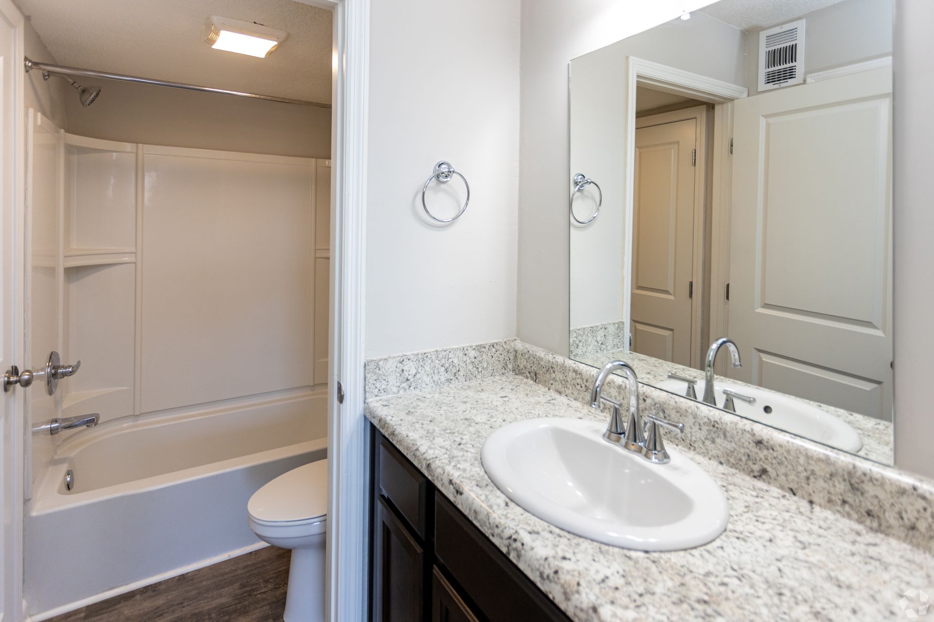 A modern bathroom with a granite vanity, single sink, dark wood cabinets, and a bathtub with a white surround.