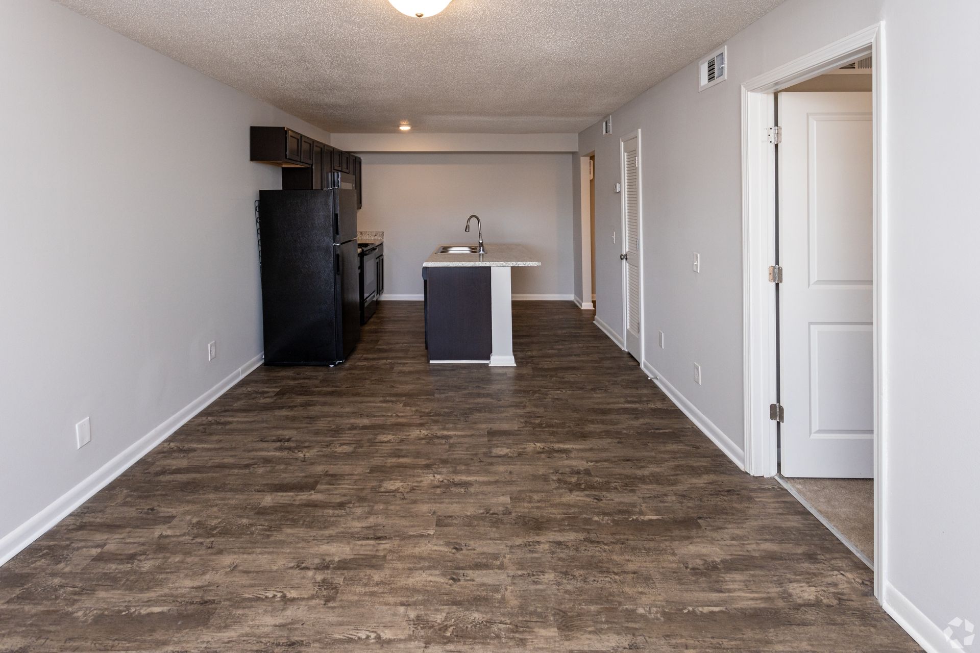 An open-concept living area with wood-look floors, featuring a kitchen island and a black refrigerator at the far end.