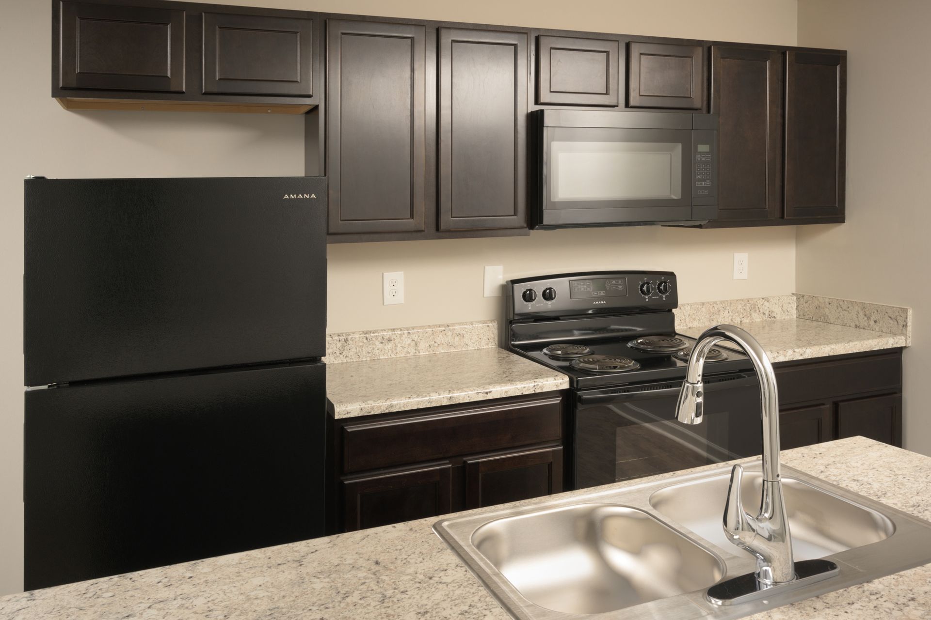 Modern kitchen with dark cabinets, black appliances, speckled light countertops, and a double-basin stainless steel sink.