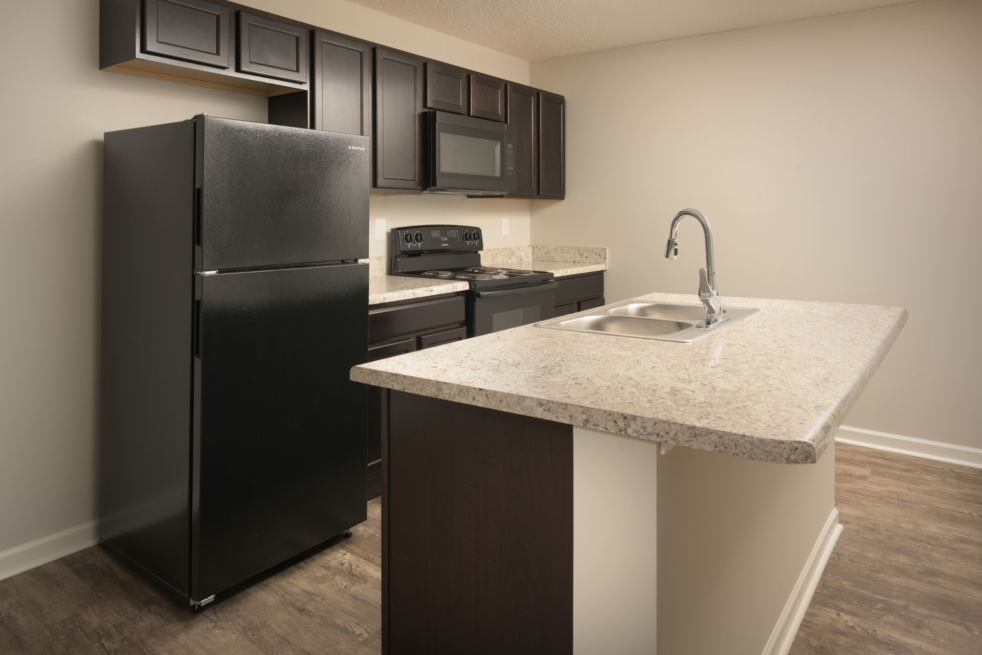 A modern kitchen with dark brown cabinets, black appliances, a granite-topped island, and light wood flooring.