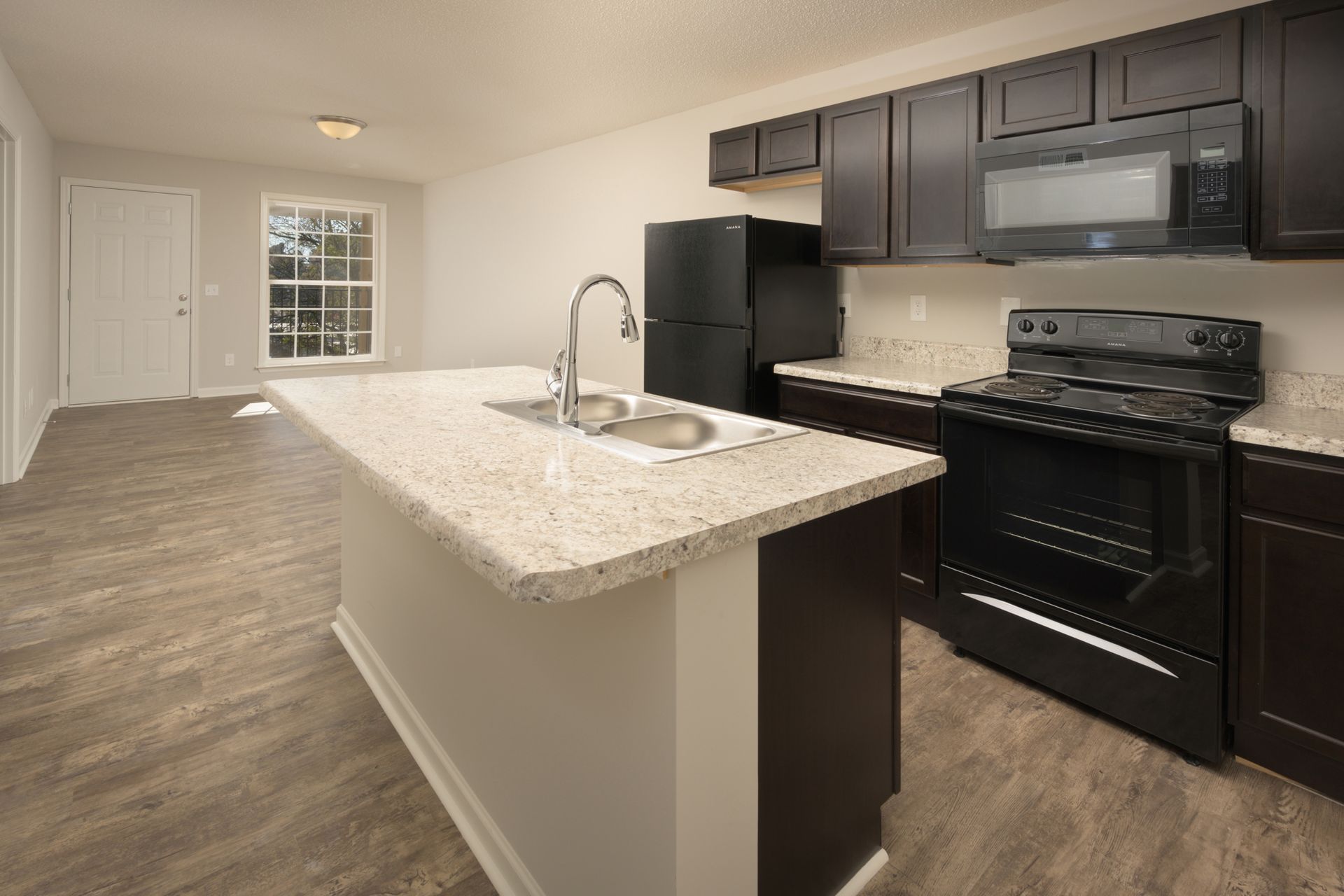 A modern kitchen featuring dark brown cabinetry, black appliances, a speckled stone-topped island, and light wood flooring.