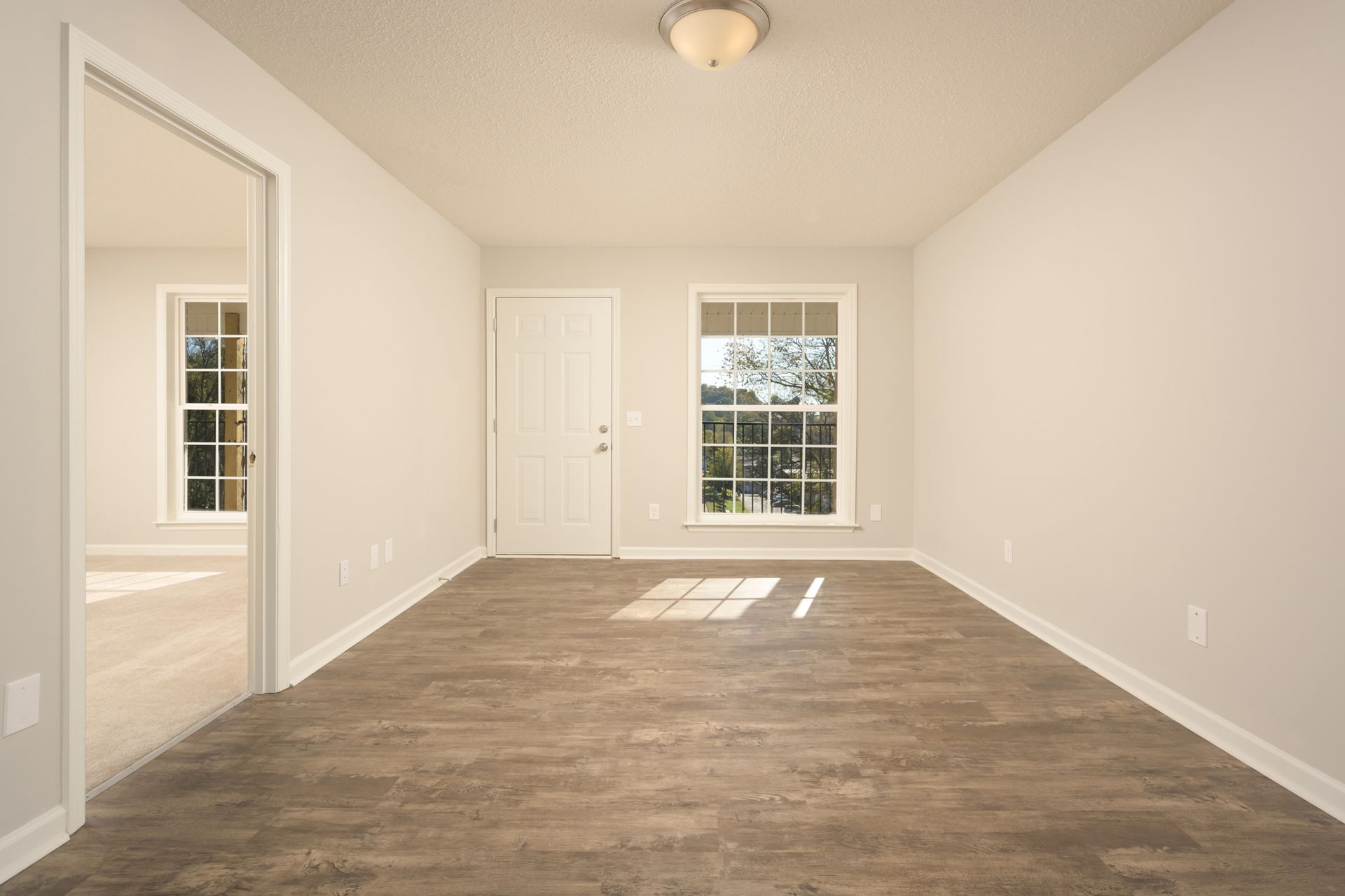 An empty room with light gray walls, brown wood-look flooring, a white door, a window, and an open doorway to another room.