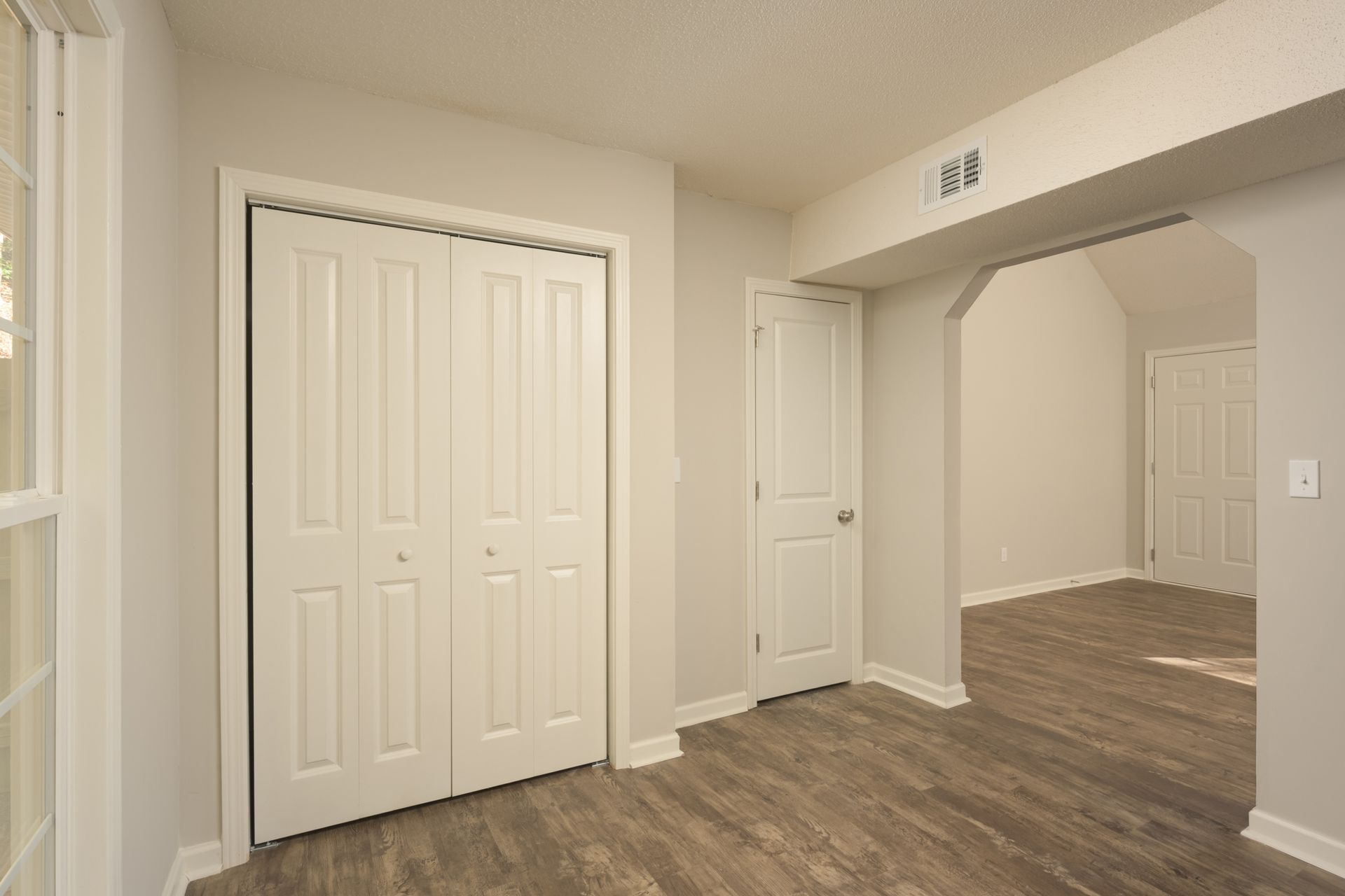 A room with light grey walls, white doors, and wood-patterned flooring, showing an open doorway to another room.