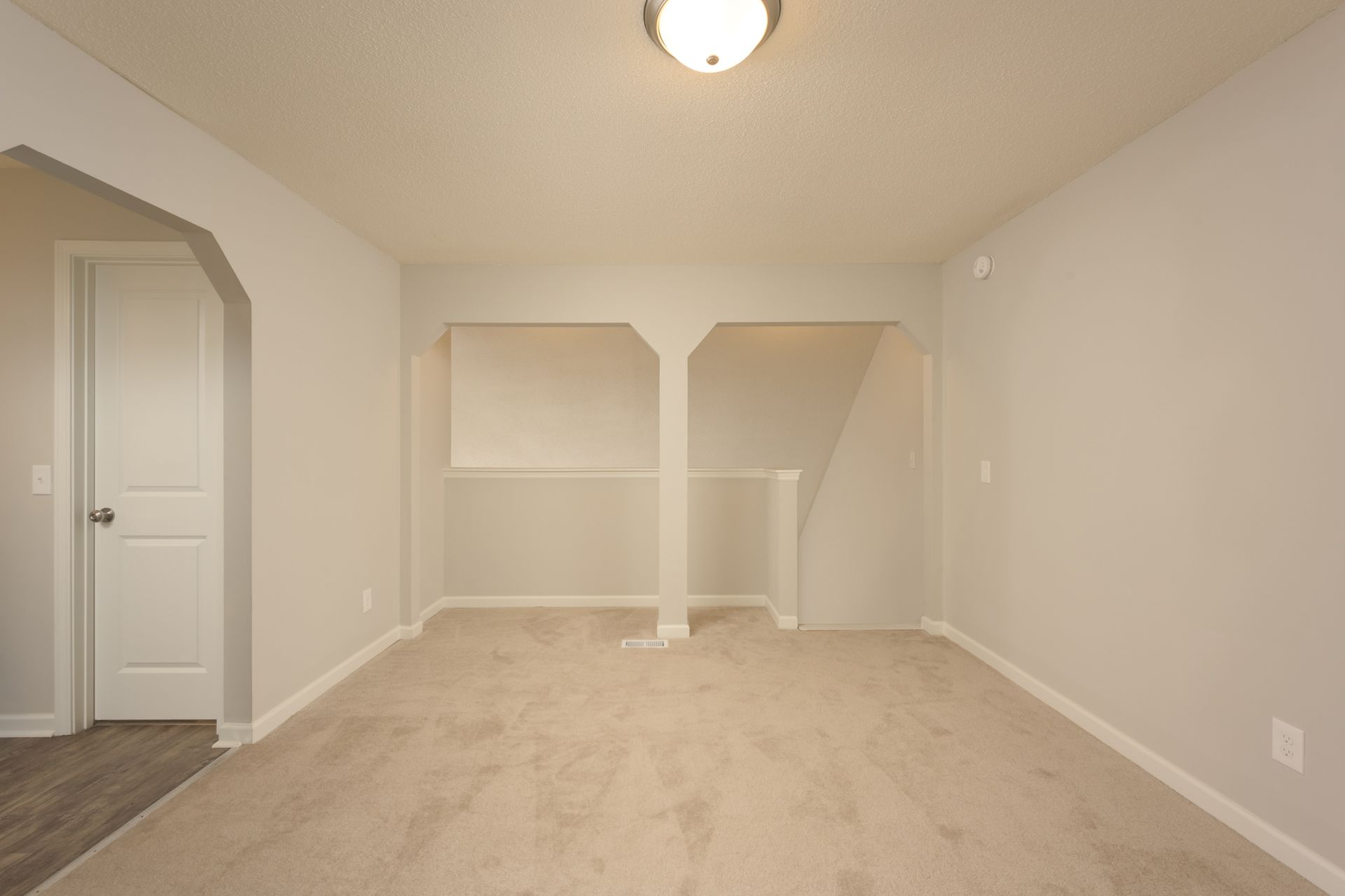 Empty room with light gray walls, tan carpet, a ceiling light, a closed white door, and an open stairwell cutout.