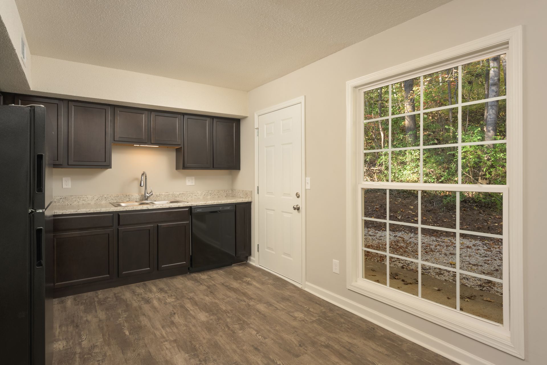 A kitchen featuring dark wood cabinets, black appliances, light-colored countertops, and a window with a wooded view.