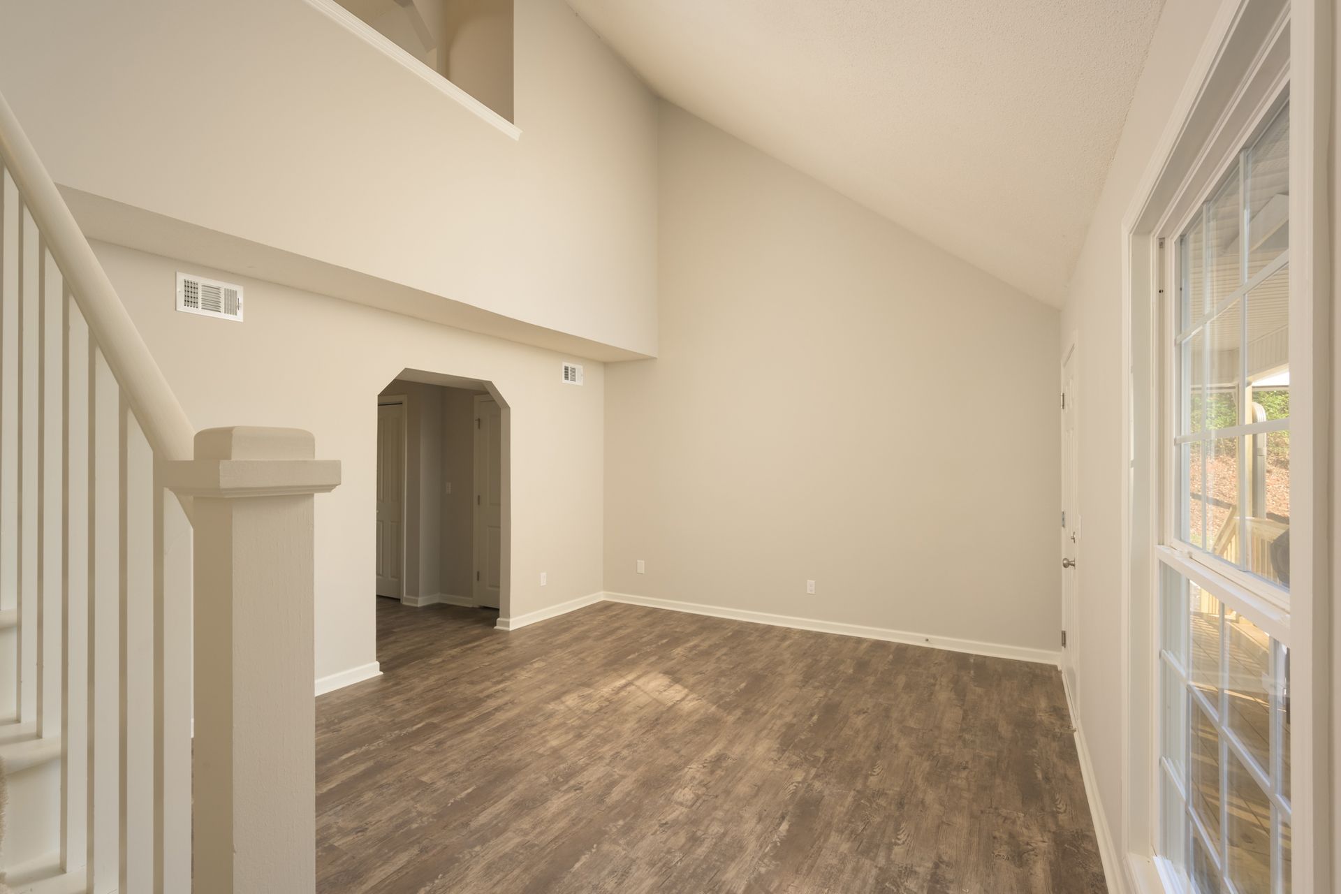 Empty room with beige walls, a high vaulted ceiling, wood-look flooring, a white staircase, and a window on the right.