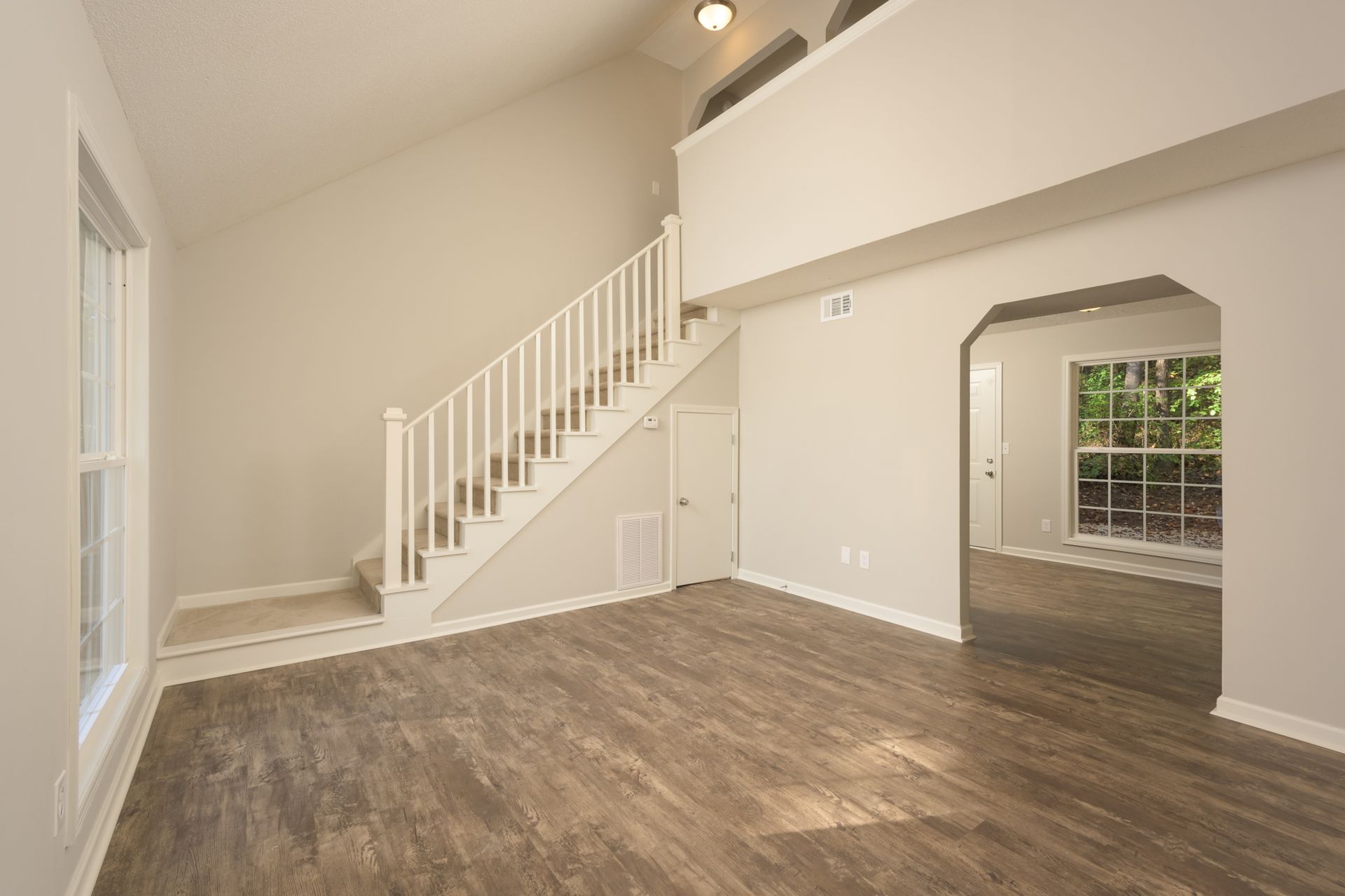 An empty room with wood-look flooring, beige walls, a white staircase, and an arched entryway to another room.