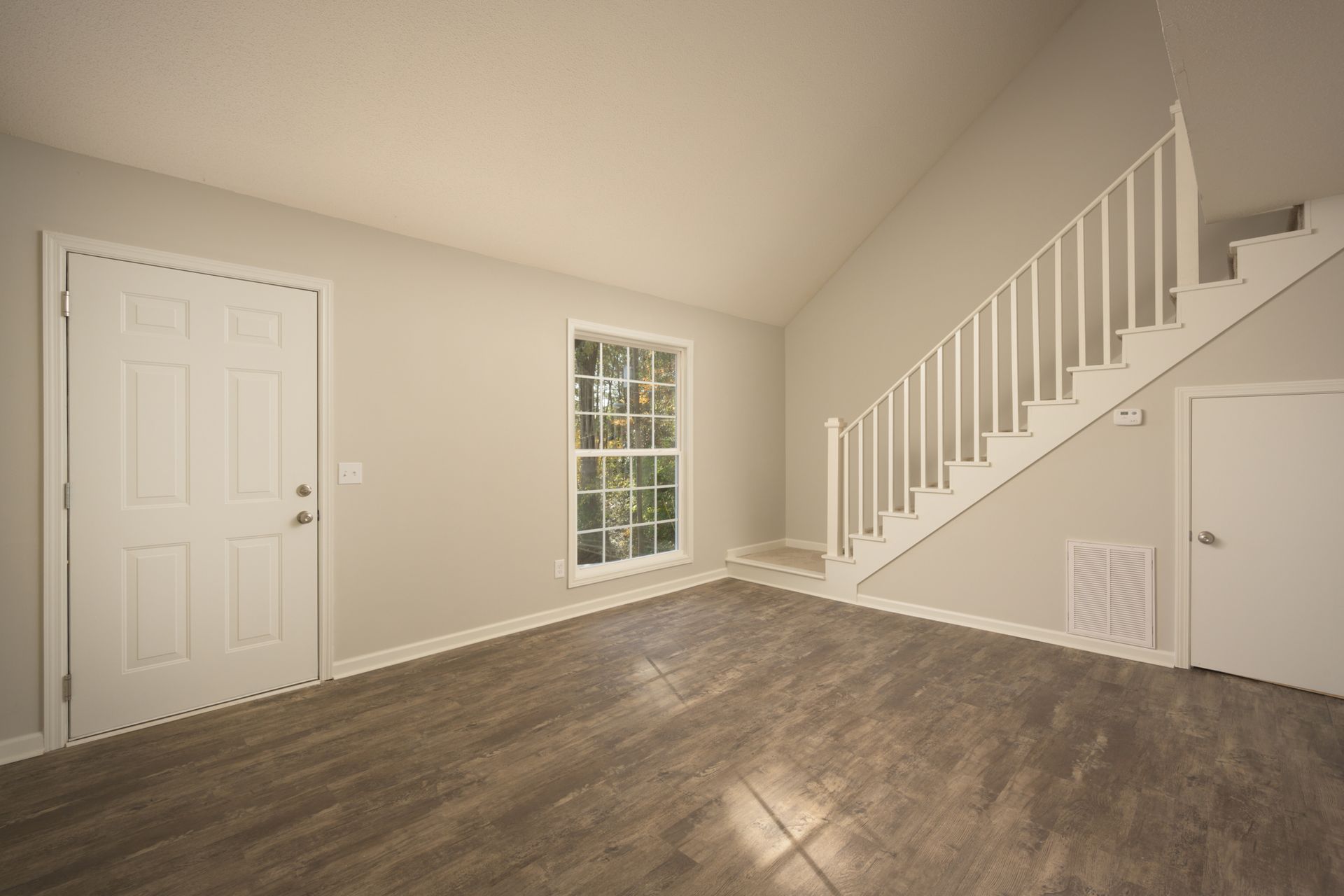 An empty room with light gray walls, wood-look flooring, a front door, a window, and a white staircase.