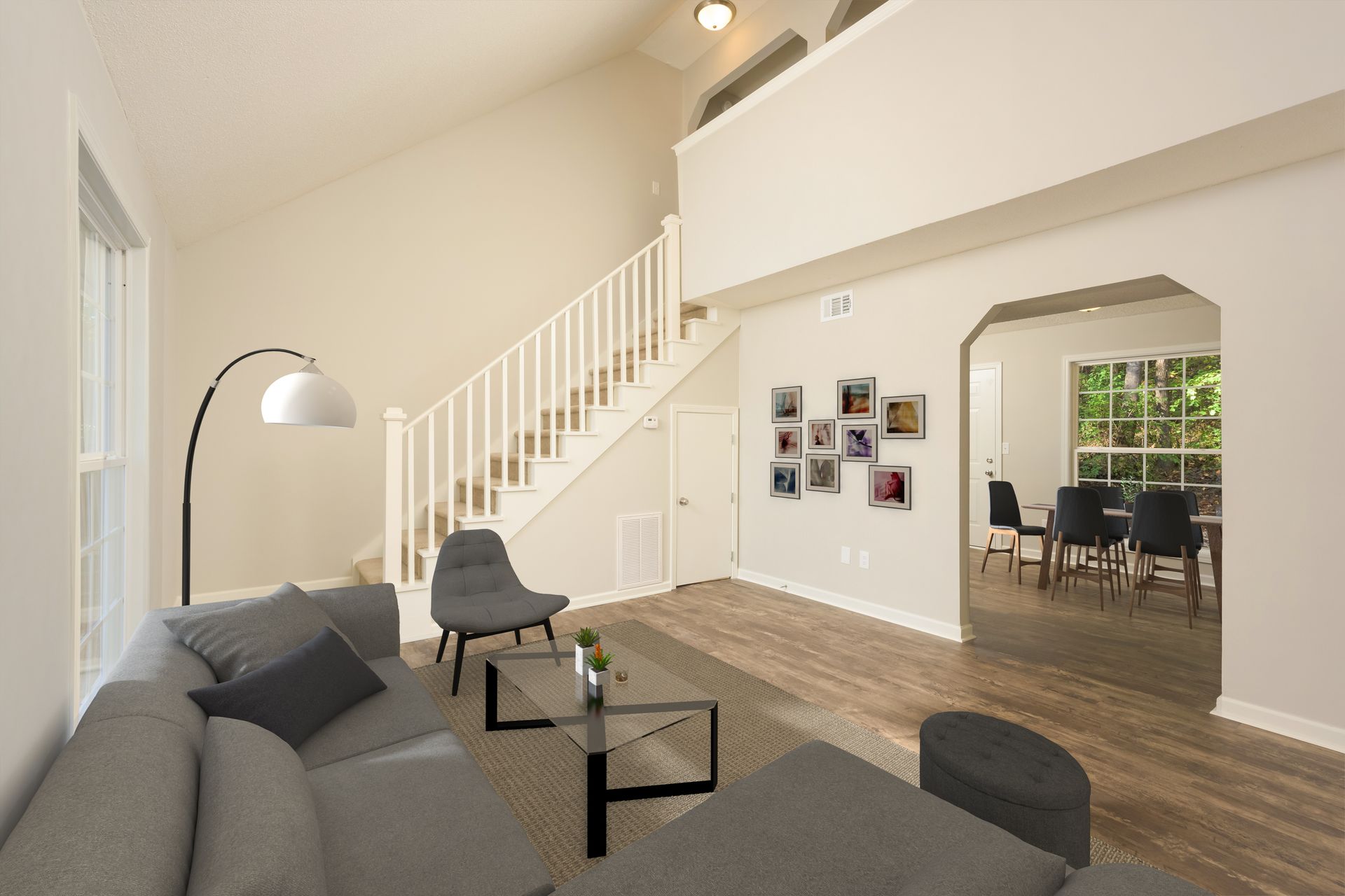 A modern living room with a grey sectional, glass coffee table, and an open stairway, opening into a dining area.