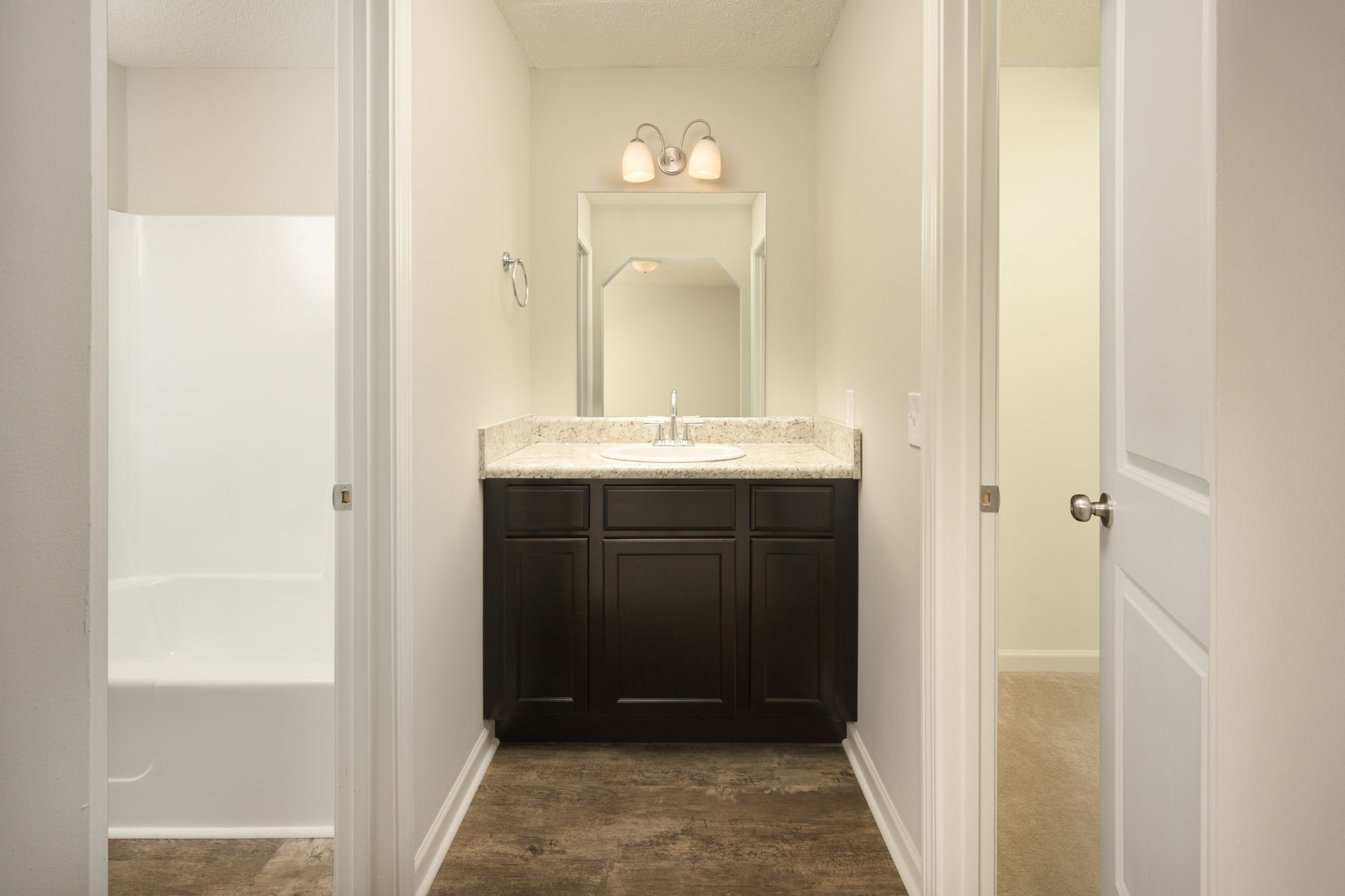 A bathroom featuring a dark wood vanity with a white countertop and mirror, flanked by a bathtub and a doorway.