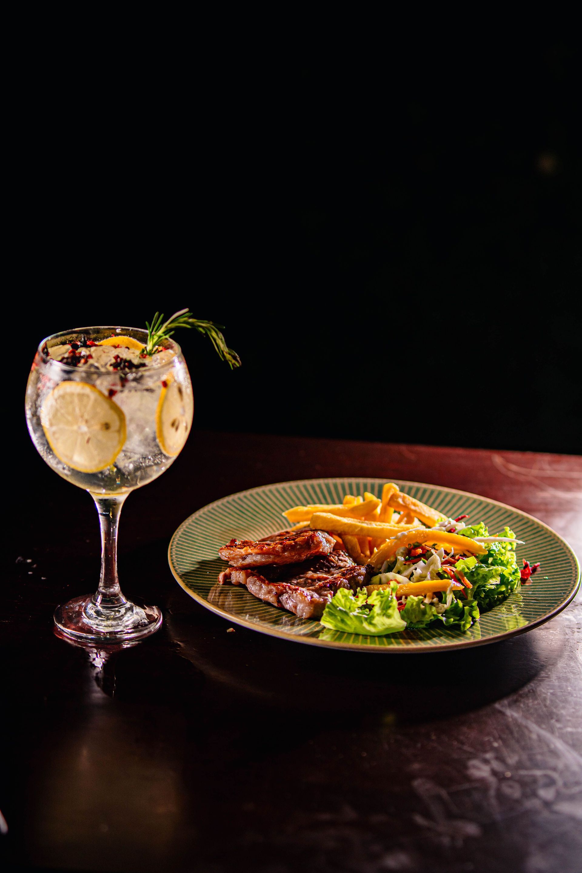 Ein Cocktail neben einem Teller mit Pommes frites, gegrilltem Fleisch und Salat auf einem dunklen Tisch.