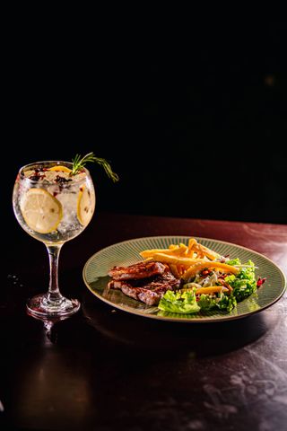 Ein Cocktail neben einem Teller mit Pommes frites, gegrilltem Fleisch und Salat auf einem dunklen Tisch.