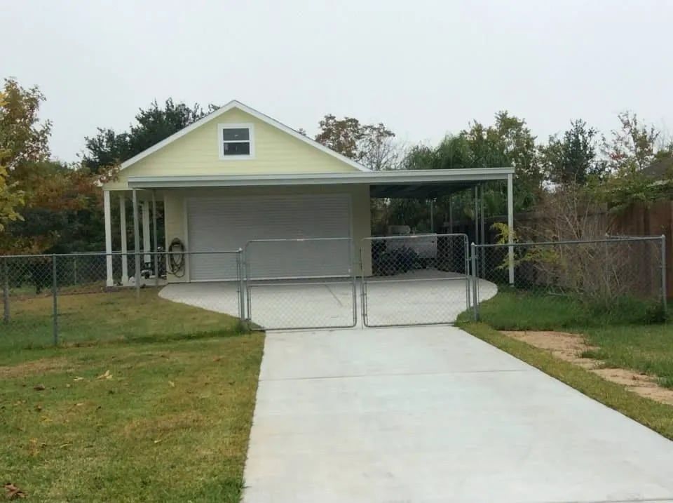 Yellow garage with carport and driveway, enclosed by chain link fence.