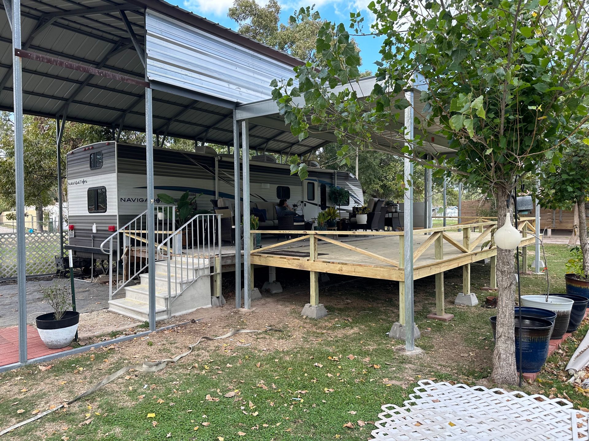 RV parked under a metal carport with a wooden deck; trees and potted plants in a grassy area.