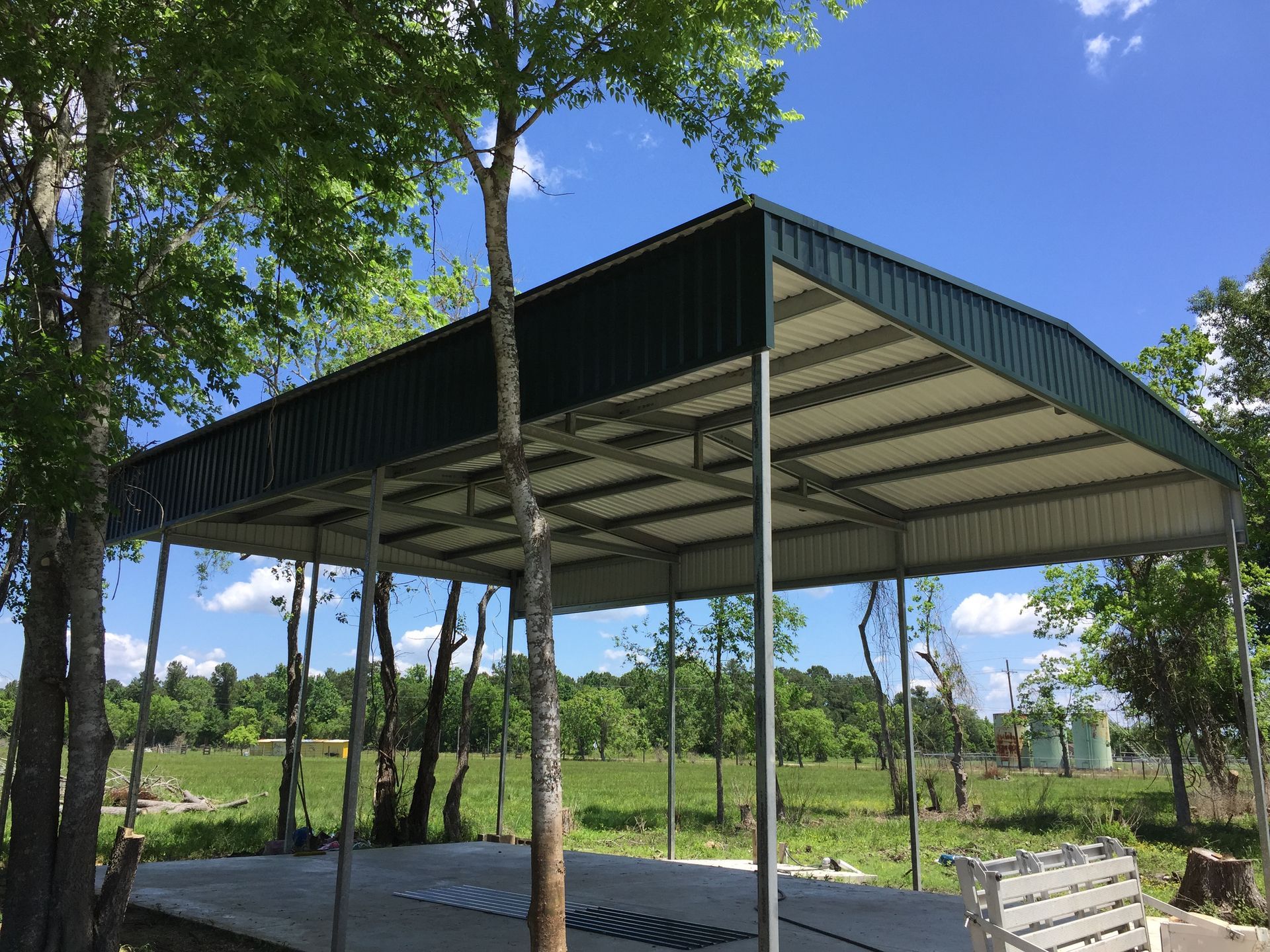Green metal roofed open-air shelter with silver support beams over a concrete pad, blue sky background.