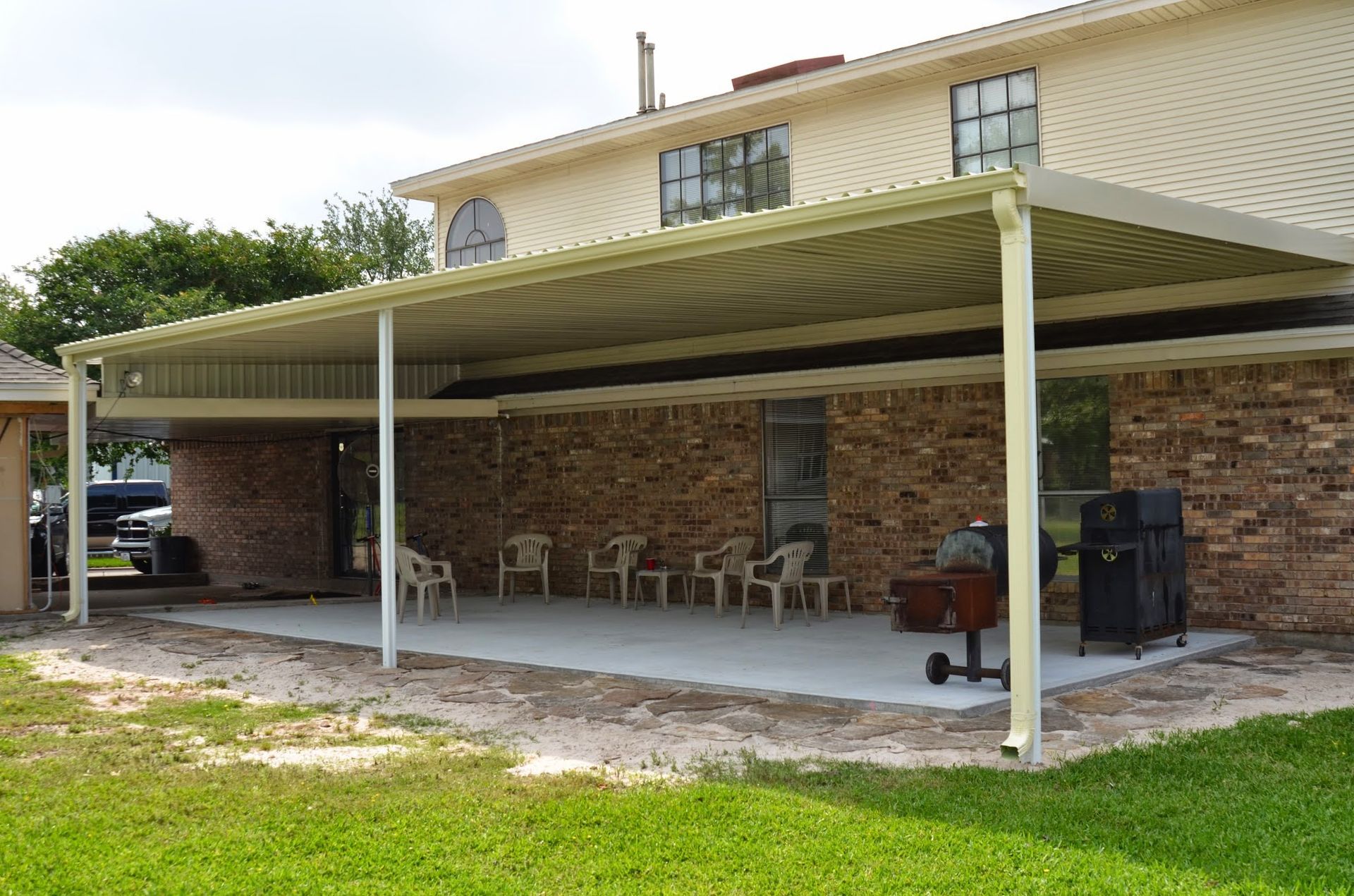 A covered patio attached to a brick house with chairs and a grill on a concrete pad.