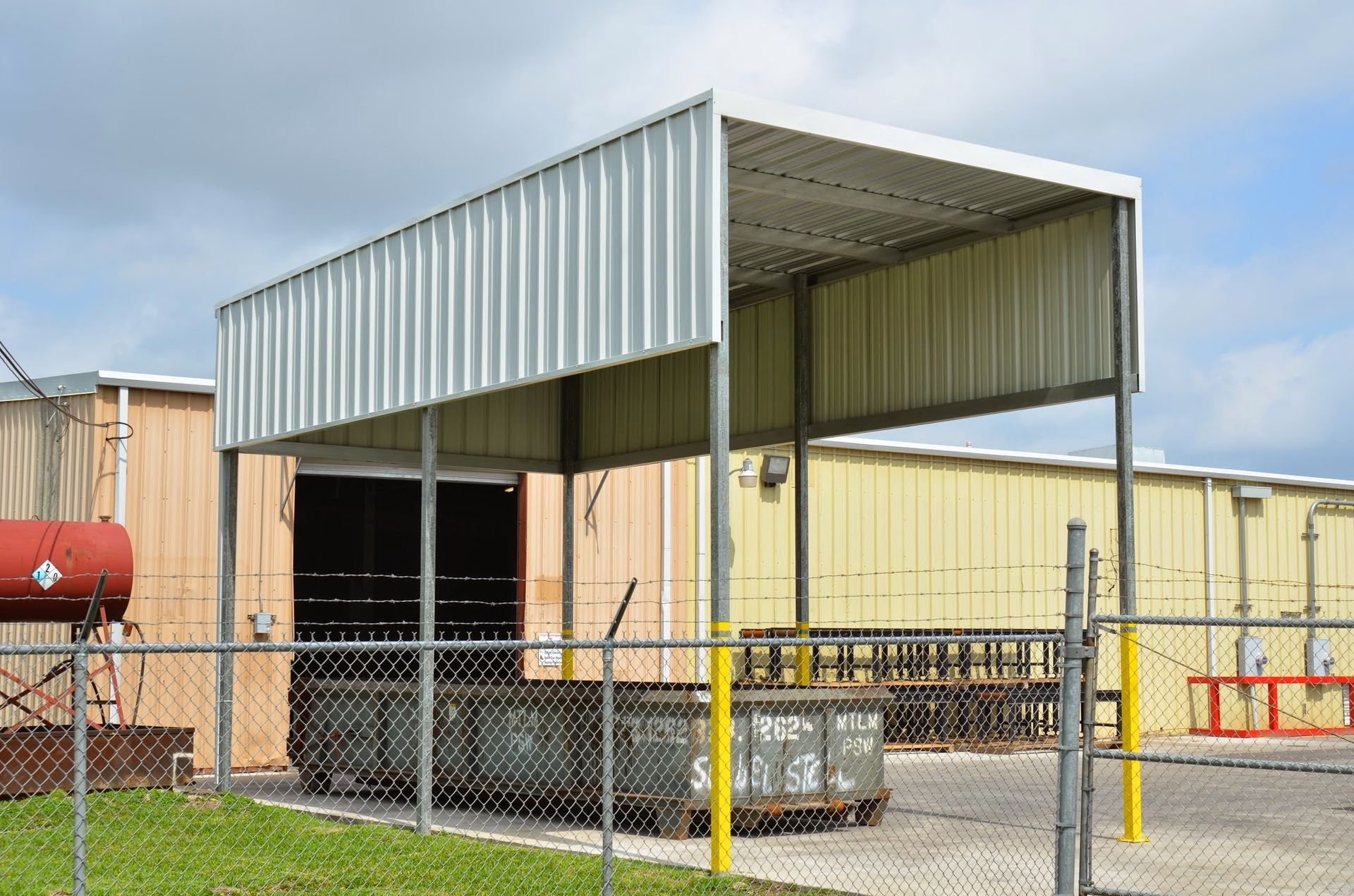 Metal shed attached to a building, over a fenced loading area with dumpsters, against a cloudy sky.