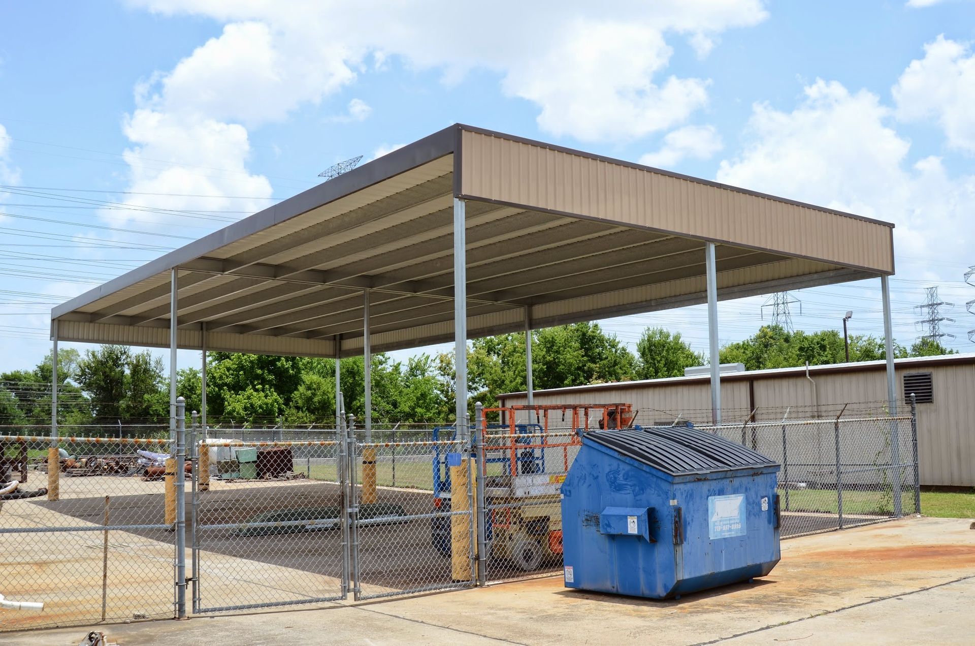 Blue dumpster under a tan metal roof with supporting poles, behind a chain-link fence.