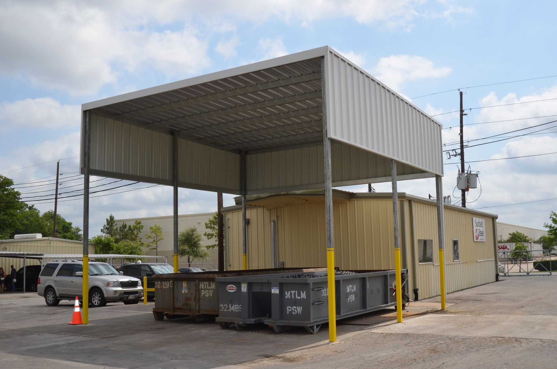 Metal shed covering two dumpsters, yellow posts, parked vehicles, overcast sky.