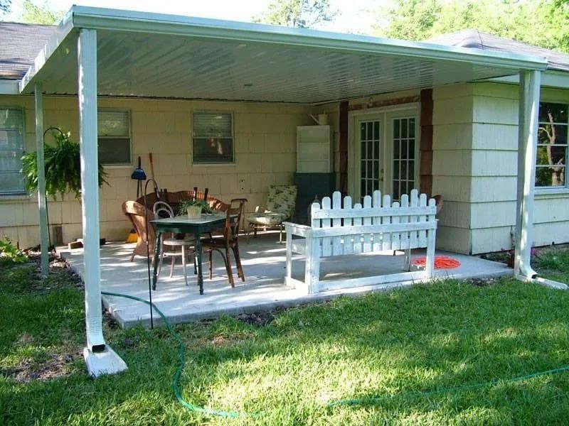 Covered patio with seating, attached to a light-colored house. Green grass and garden in front.