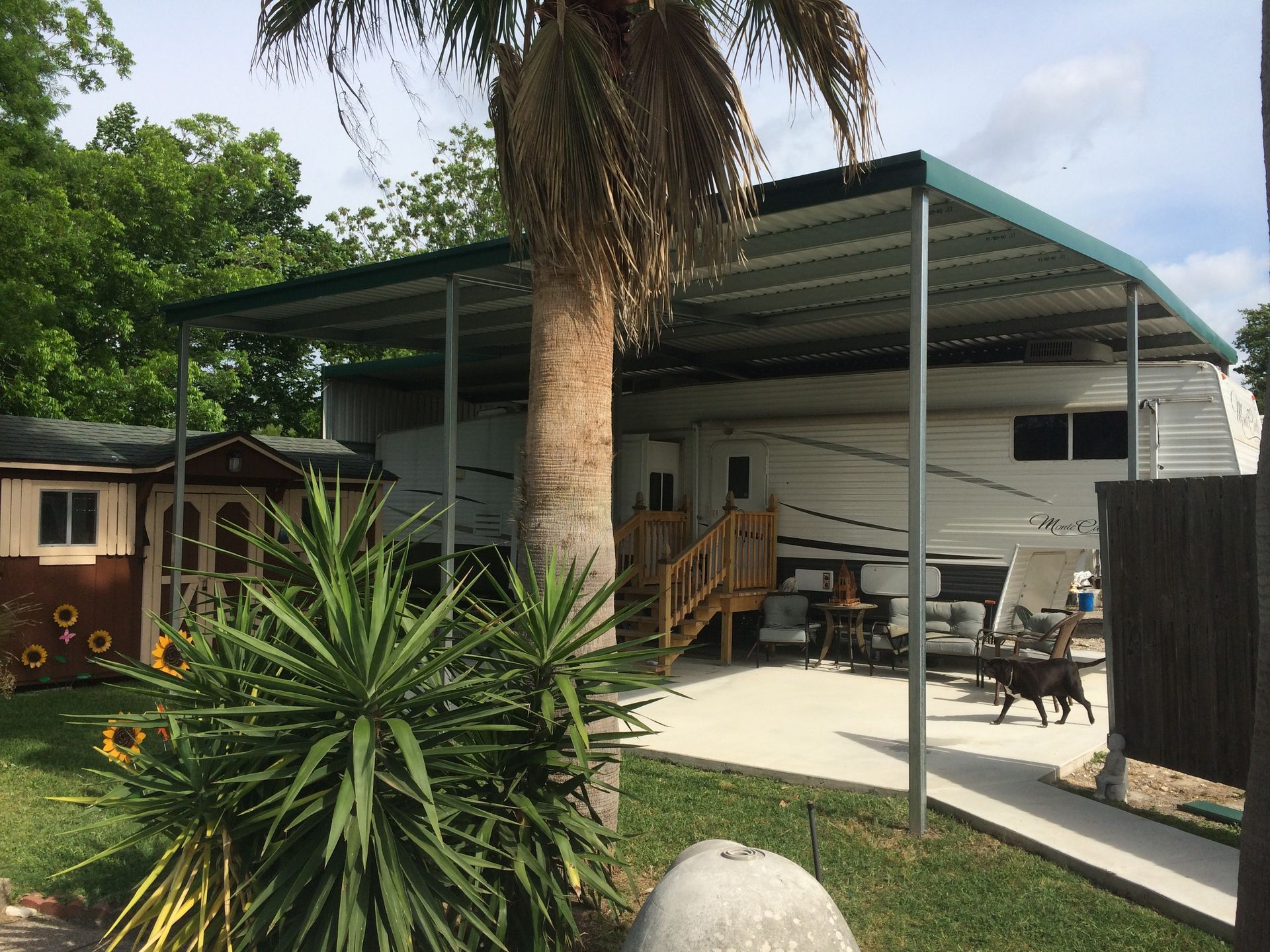 RV parked under a green metal carport, with a small dog and a palm tree in a grassy yard.