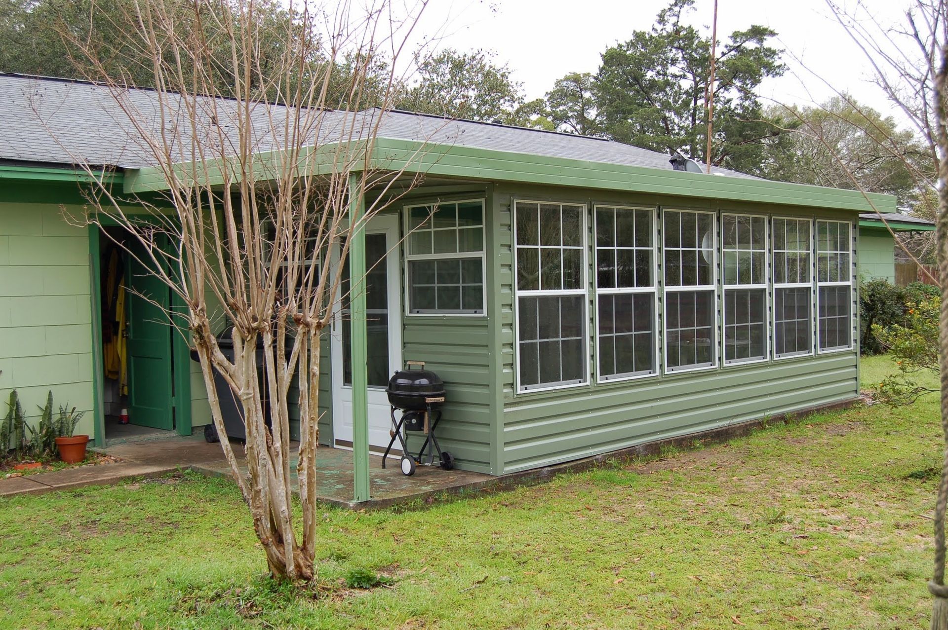 Green-sided porch with many windows attached to a green house. A grill sits outside the doorway.
