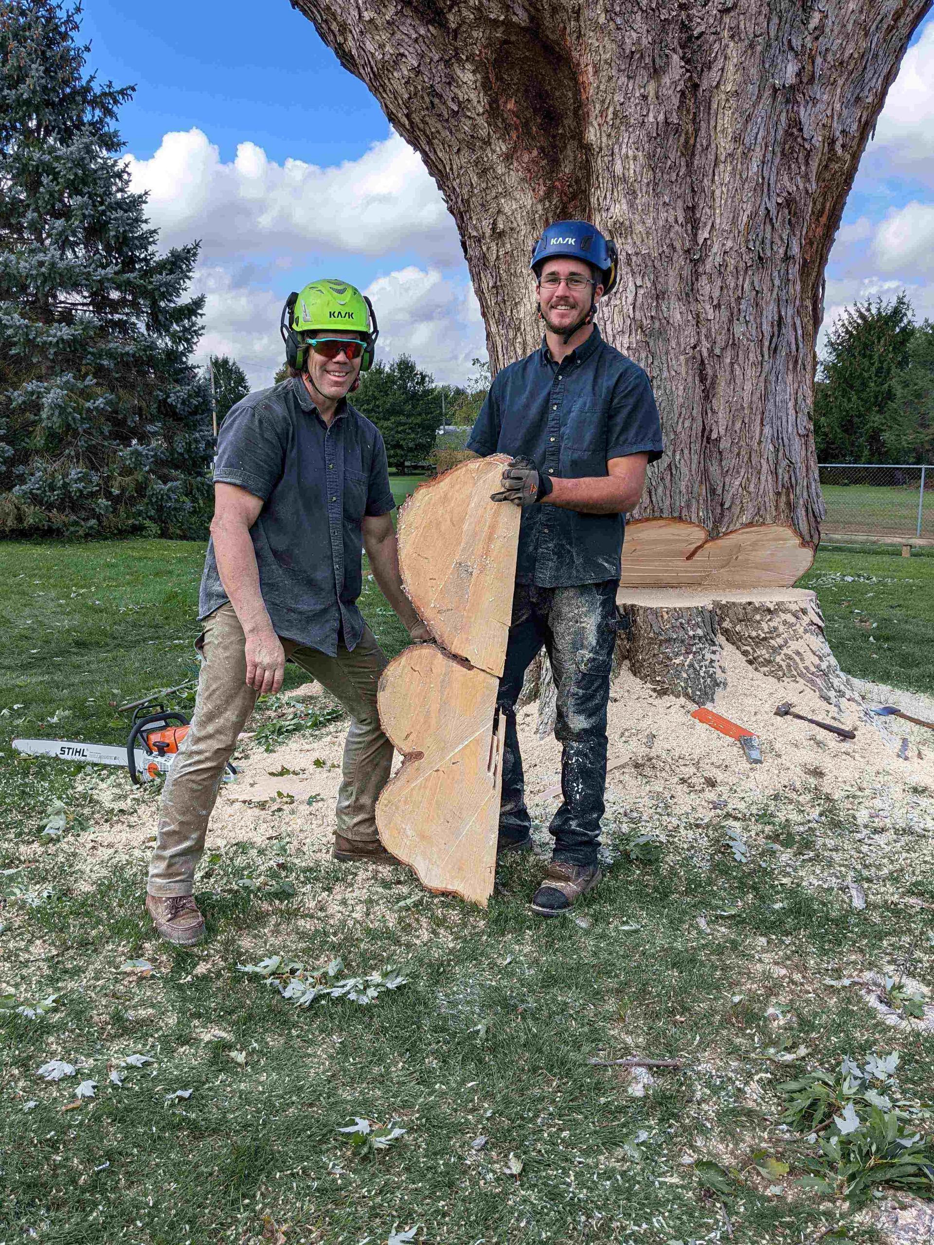 Two men are standing next to a tree stump holding a piece of wood.