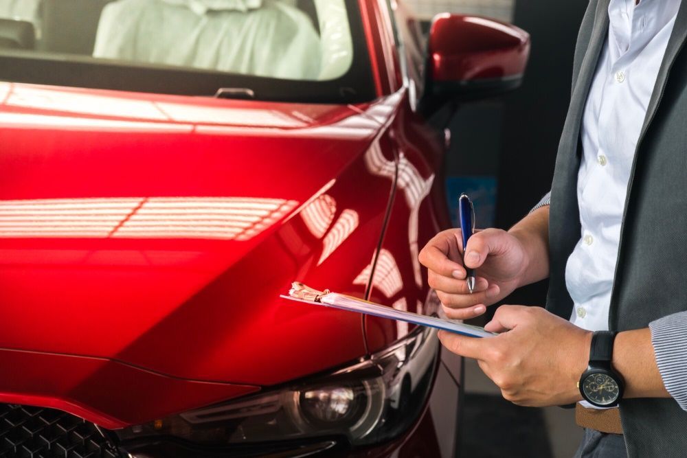 A Man is Writing on a Clipboard in Front of a Red Car — Forster Mechanical Repairs & Servicing In Forster, NSW