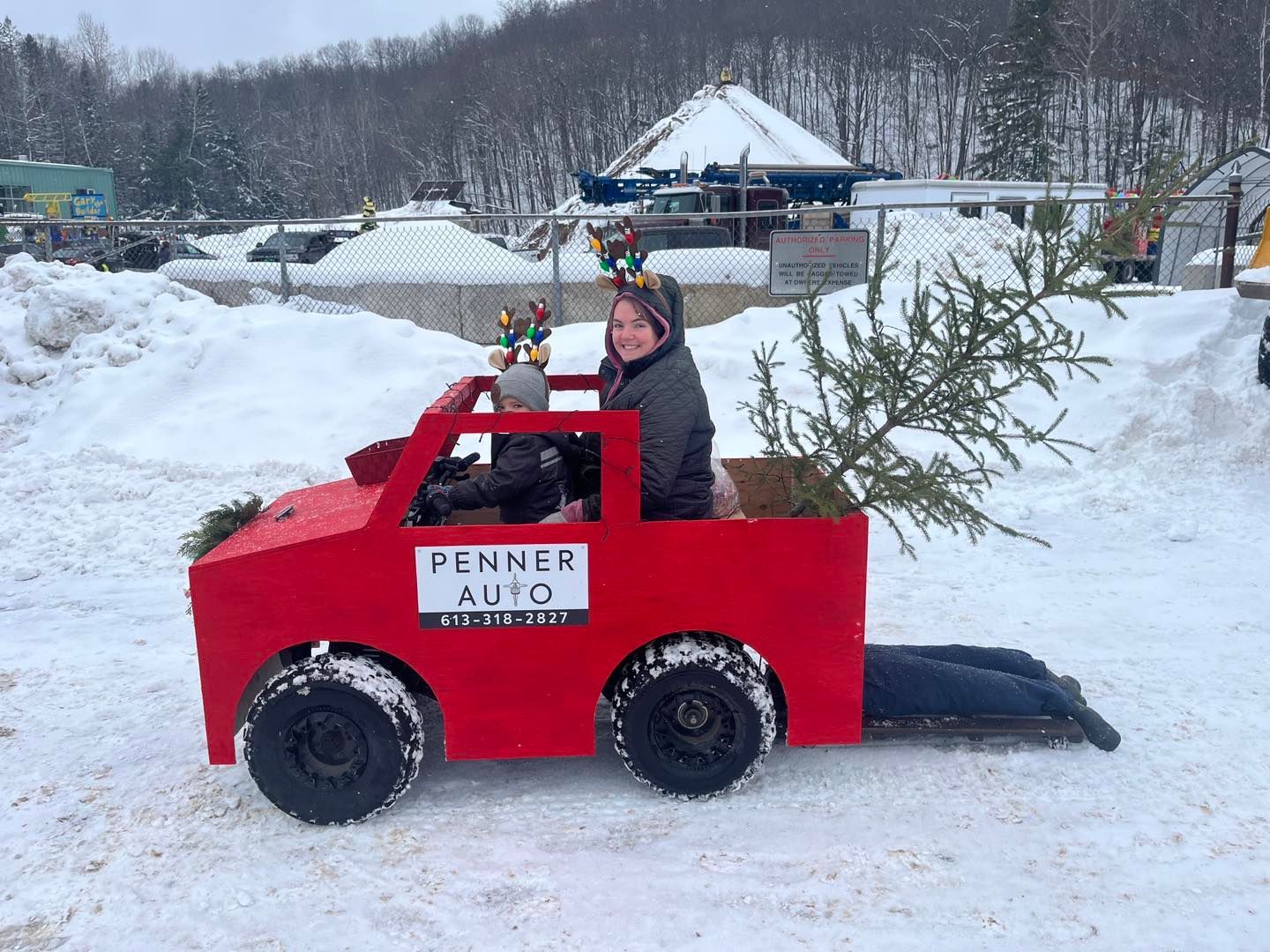 Person in a red toy car with a Christmas tree, driving in a snowy outdoor event area. | Penner Auto