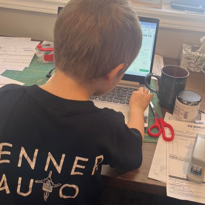 Child working at a desk with a laptop, papers, scissors, and a black mug near a window. | Penner Auto