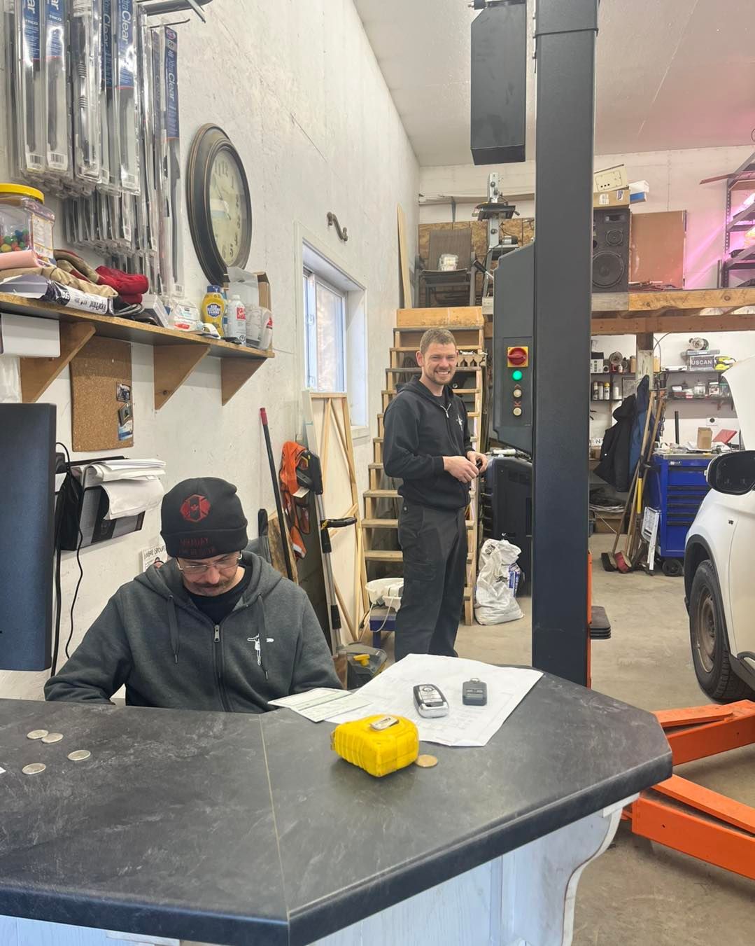 Auto repair shop with two workers, one seated at a desk and one standing near a lift in the background. | Penner Auto