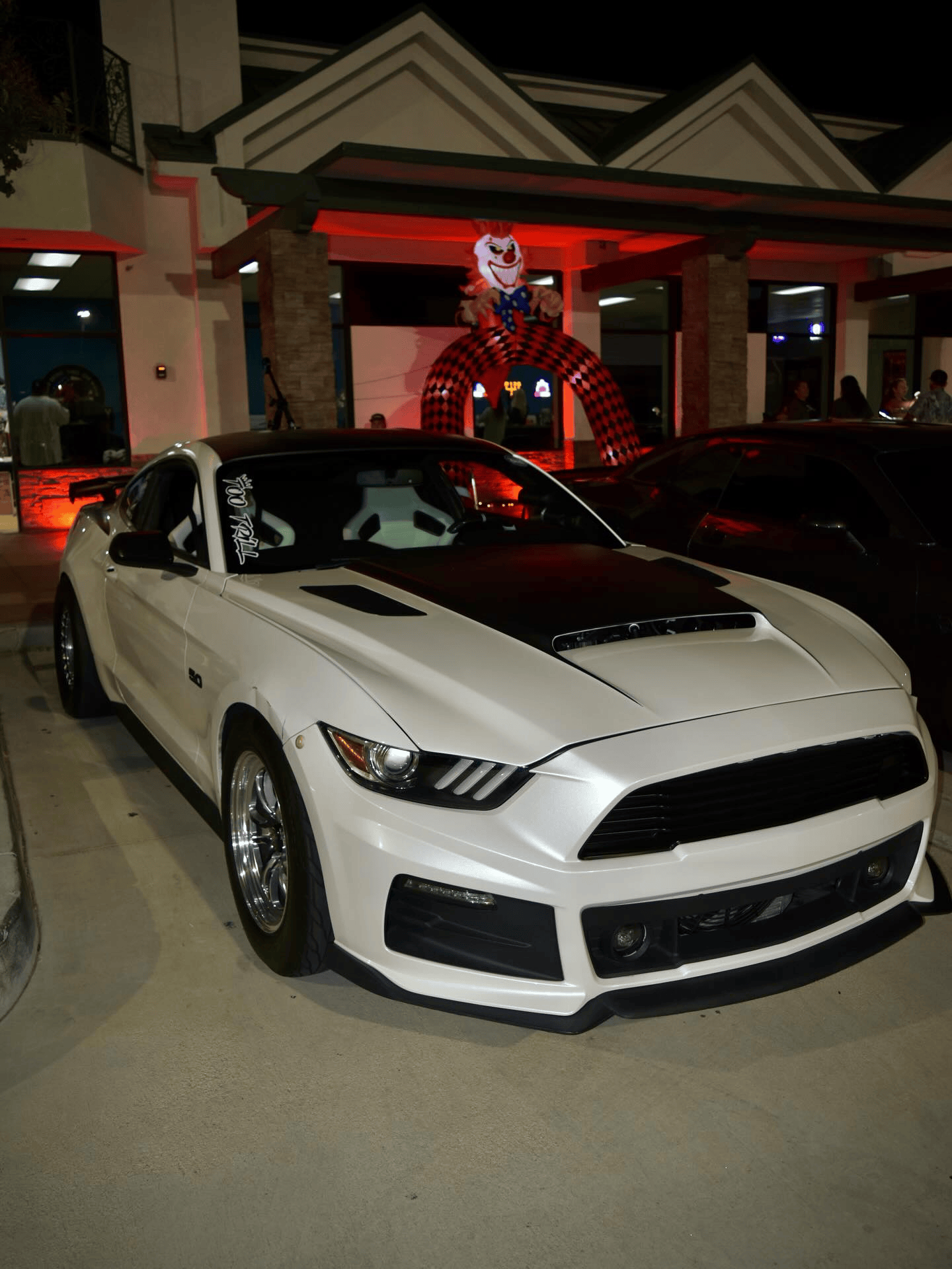 White sports car parked at a lit building entrance with a large clown figure in the background