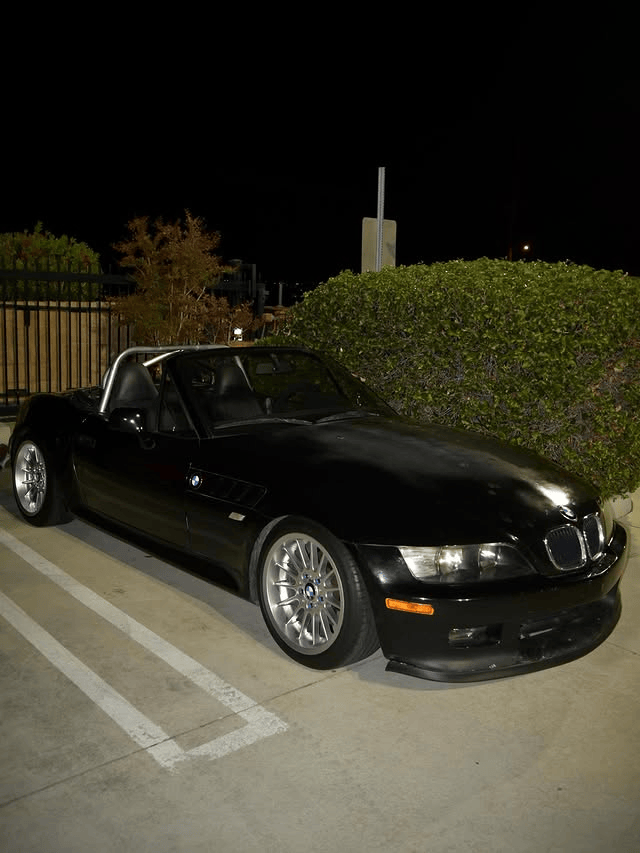 Black BMW sports car parked at night beside a hedge in a marked parking lot