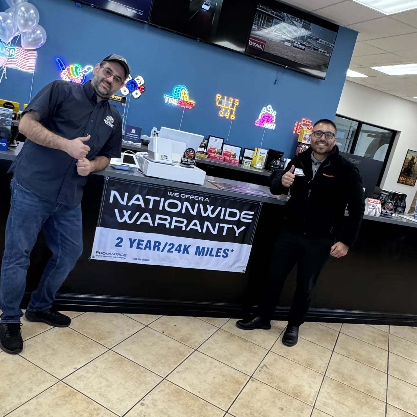 Two people posing by a Nationwide Warranty service counter, one holding a sign reading “2 YEAR/24K MILES.”