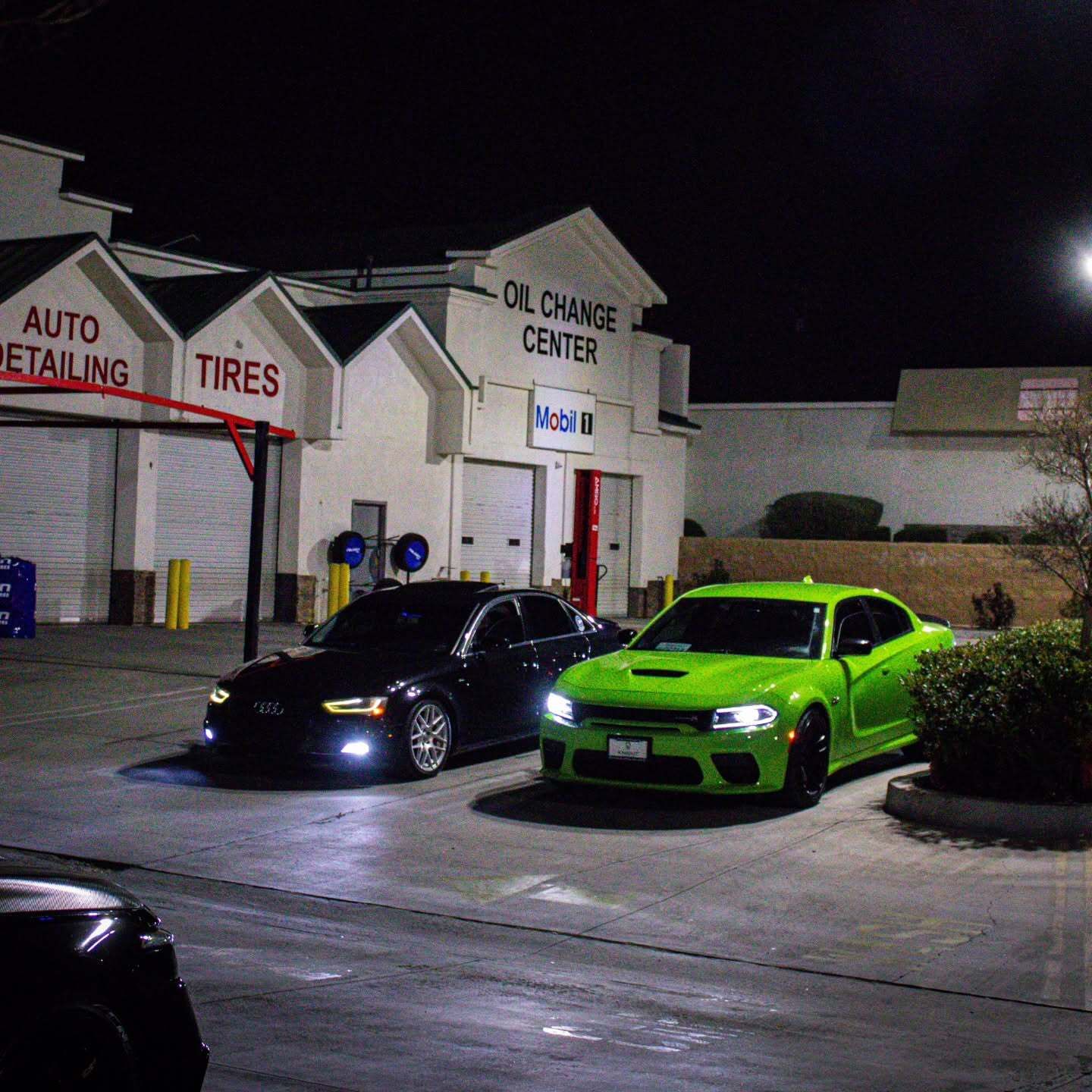 Two parked cars, black and neon green, outside an auto detailing and oil change center at night