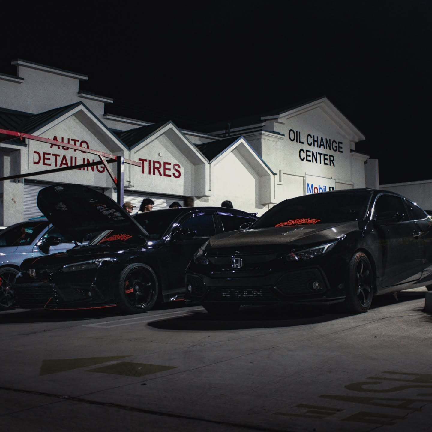 Two black sports cars parked at a nighttime auto detail and tire shop.