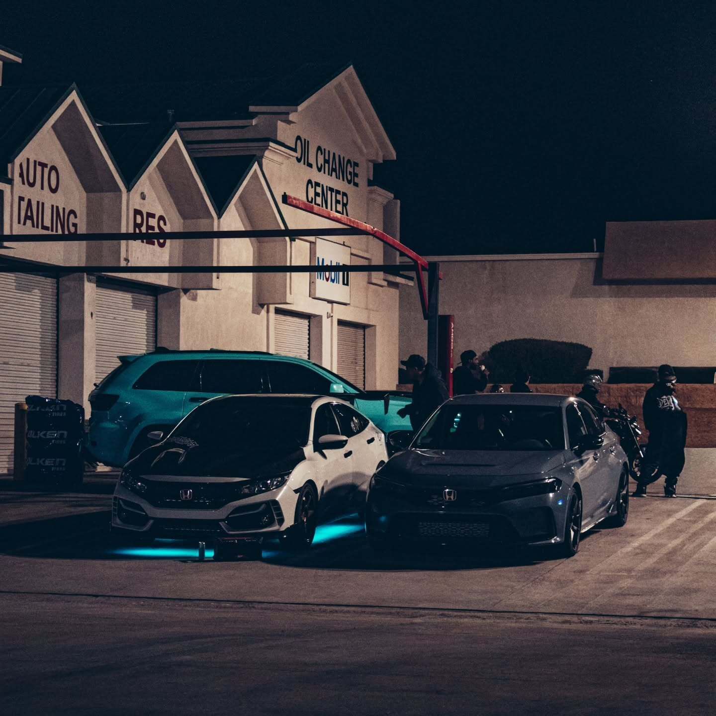 Two black cars parked at night outside a failing auto shop, with teal underglow lighting.