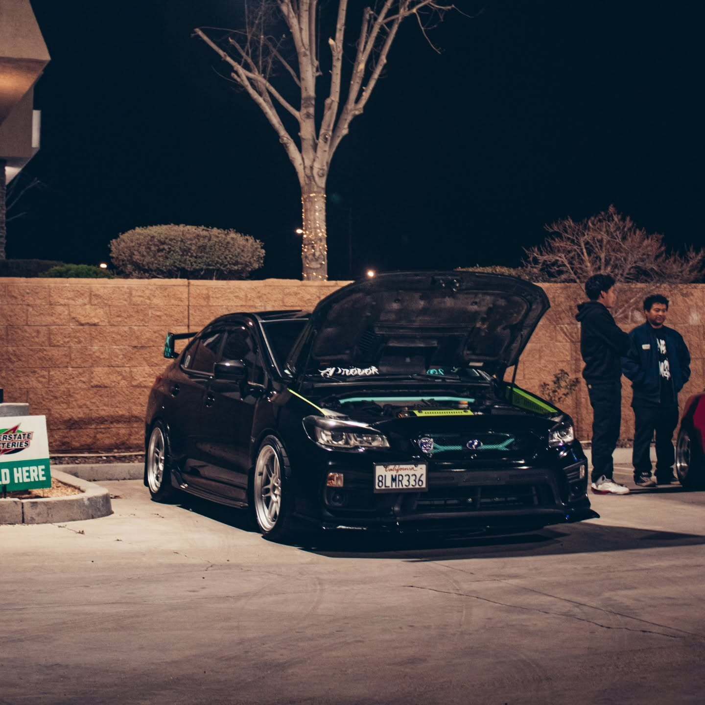 Black modified sedan with hood open at night in a parking lot, two people standing nearby