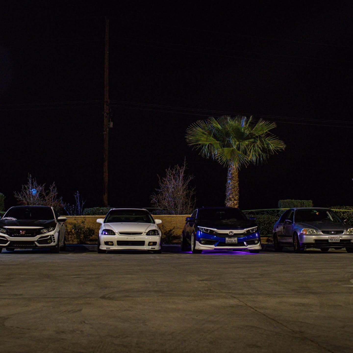 Four cars parked at night in a lot, with a palm tree and blue underglow lights on one car.