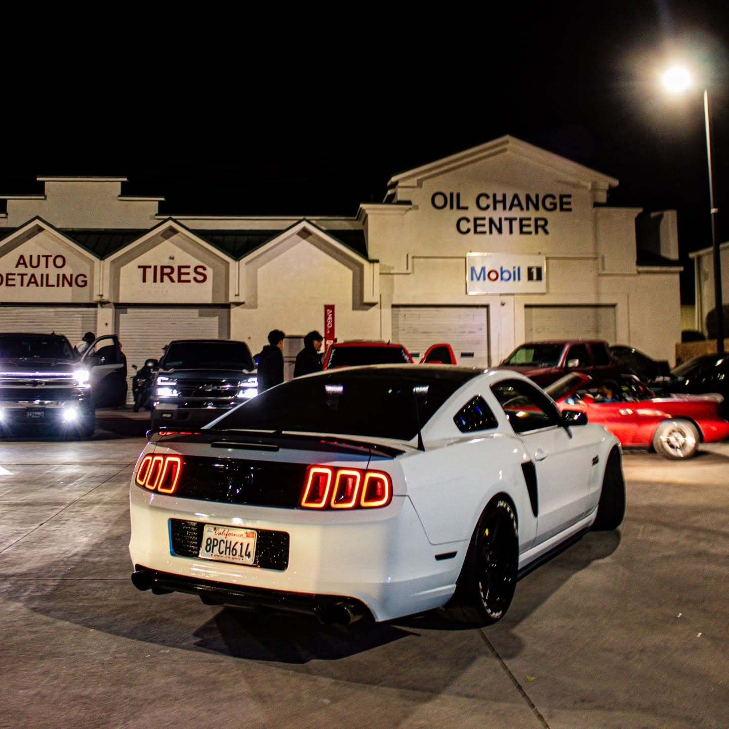 White Ford Mustang parked at night outside an auto shop, with other cars and lit signs in the background