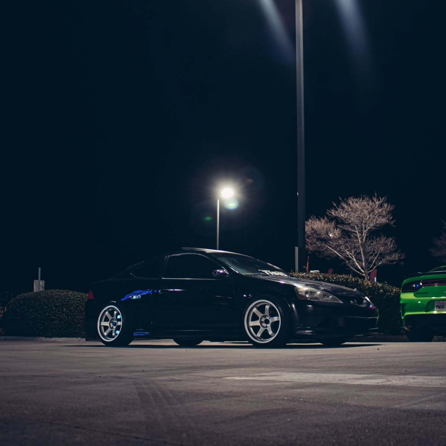 Black sports car parked at night under streetlights, with a green car in the background.